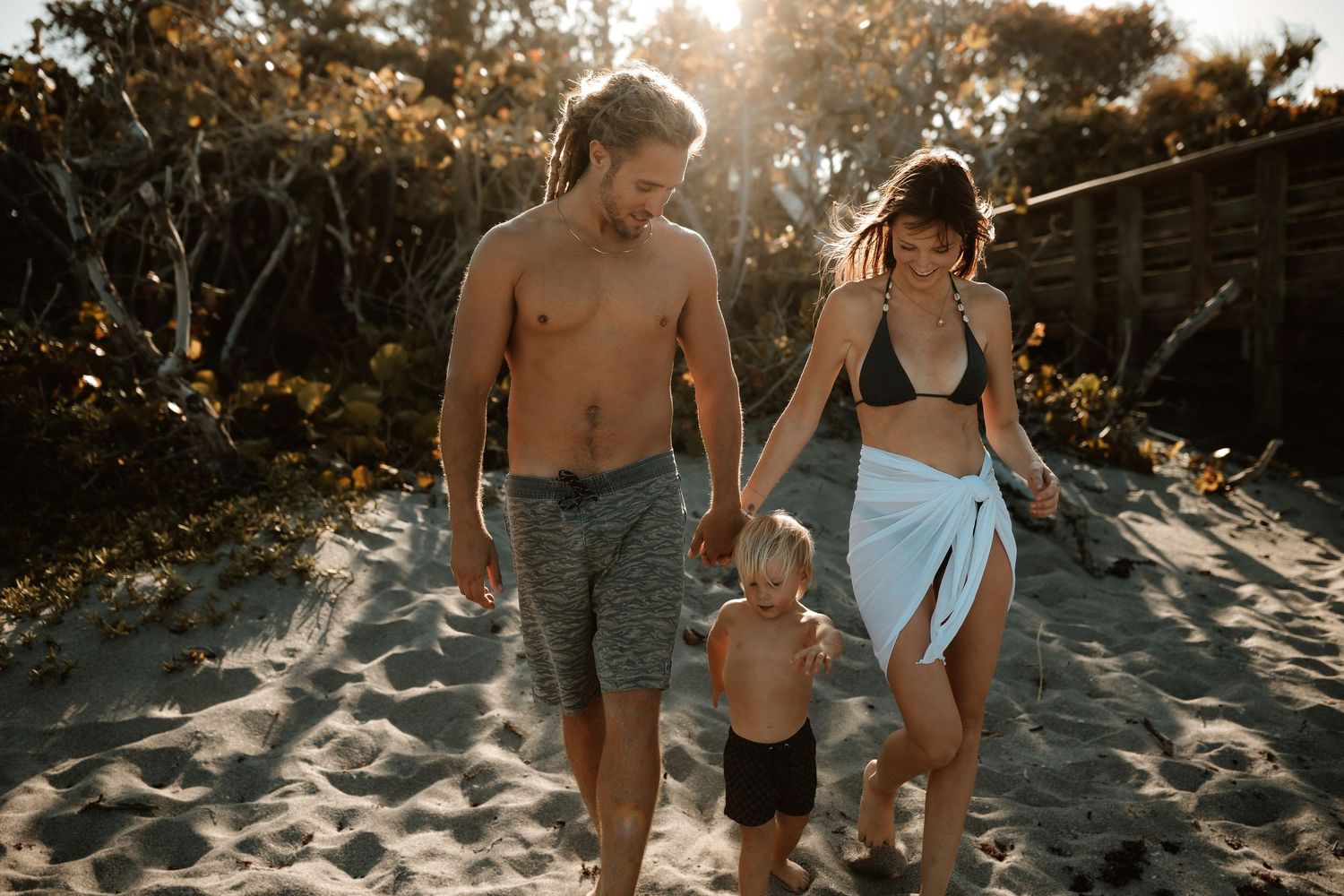Family of three walking on sandy beach path, parents holding toddler's hands in golden sunlight.