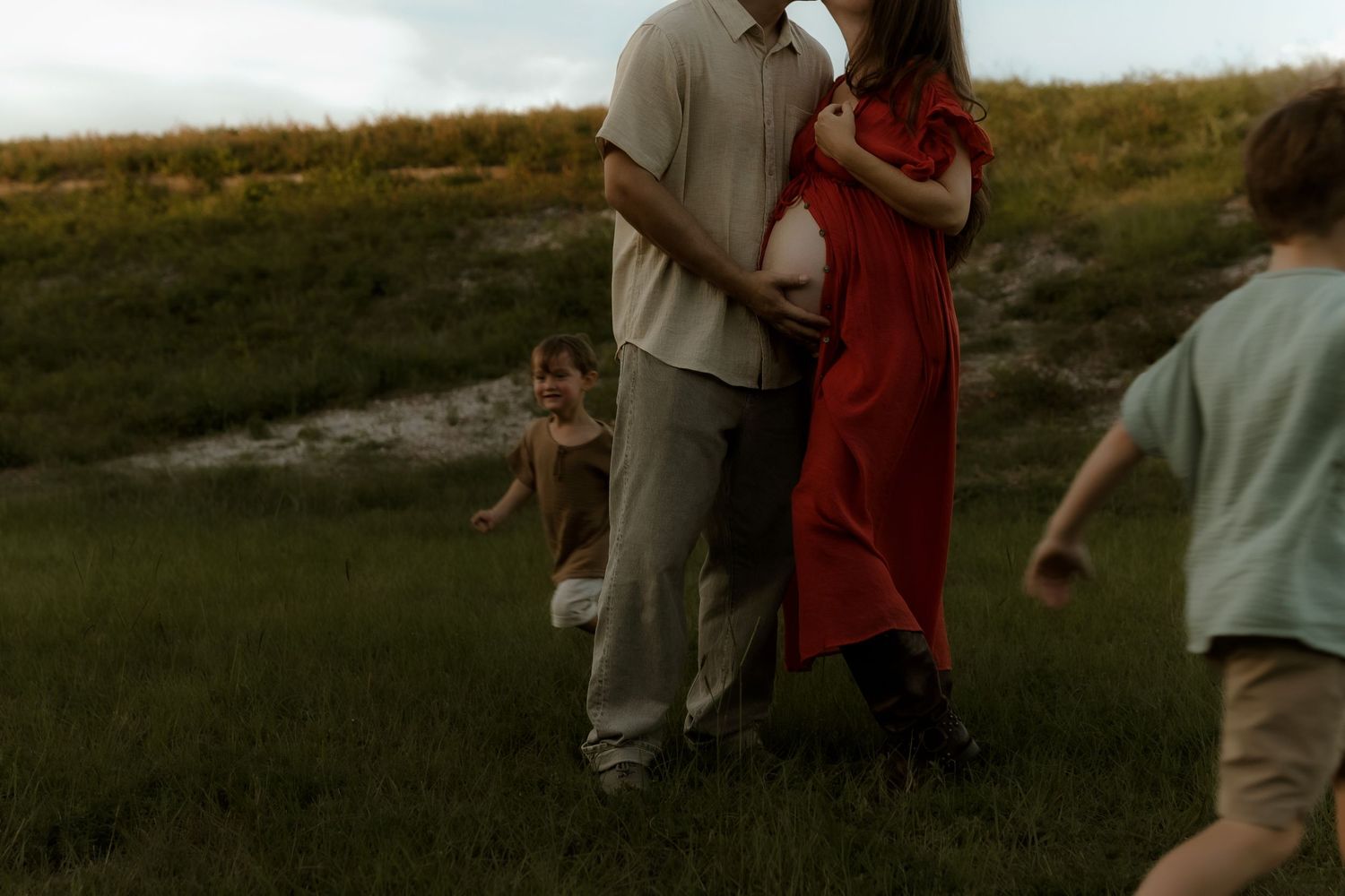 Pregnant couple embracing in green field while two young children play around them during outdoor maternity photo session.