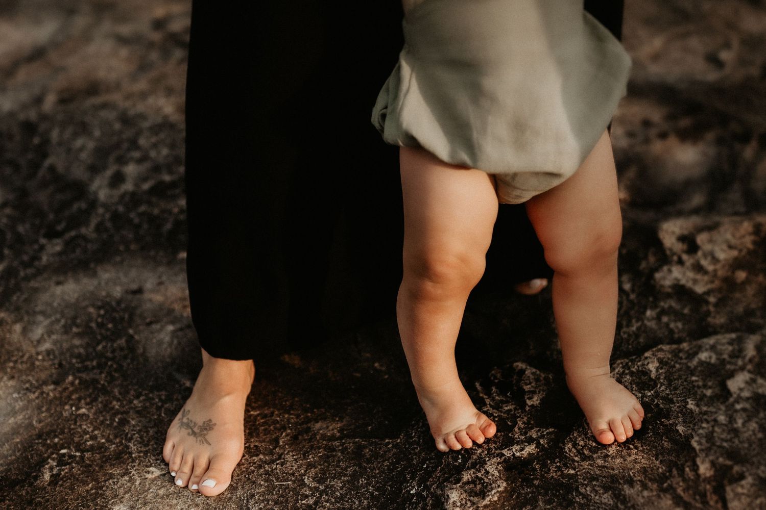 Adult and baby barefoot on rocky ground, child in sage green dress standing between parent's legs.