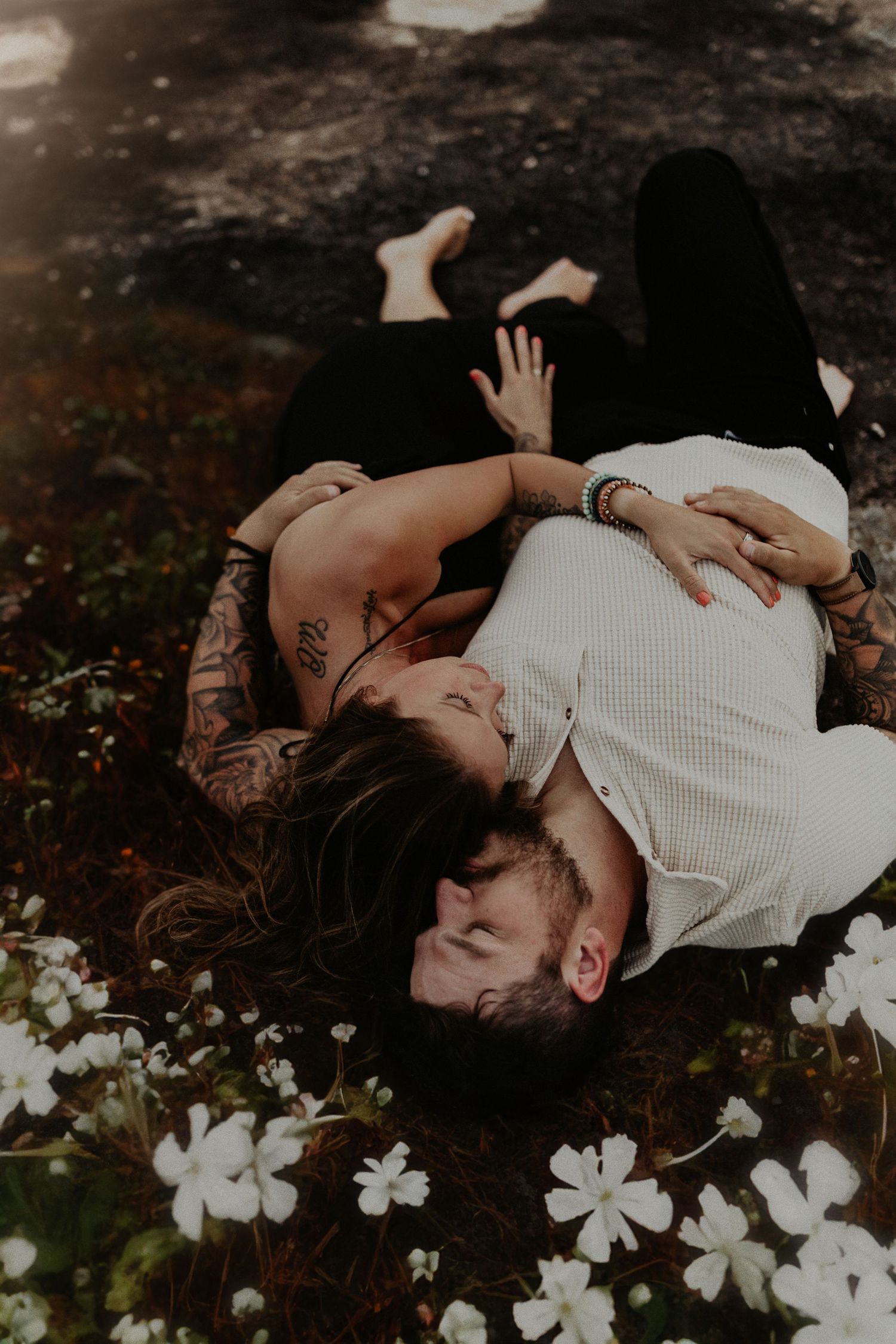 Couple lying in a field of white flowers, embracing romantically, woman with tattoos smiling at man in white shirt.