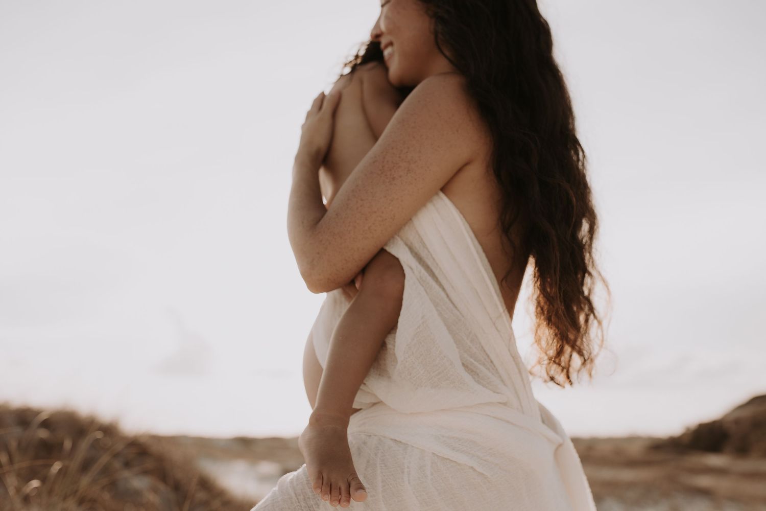 Pregnant woman in white wrap fabric holds toddler close on sandy beach at golden hour.