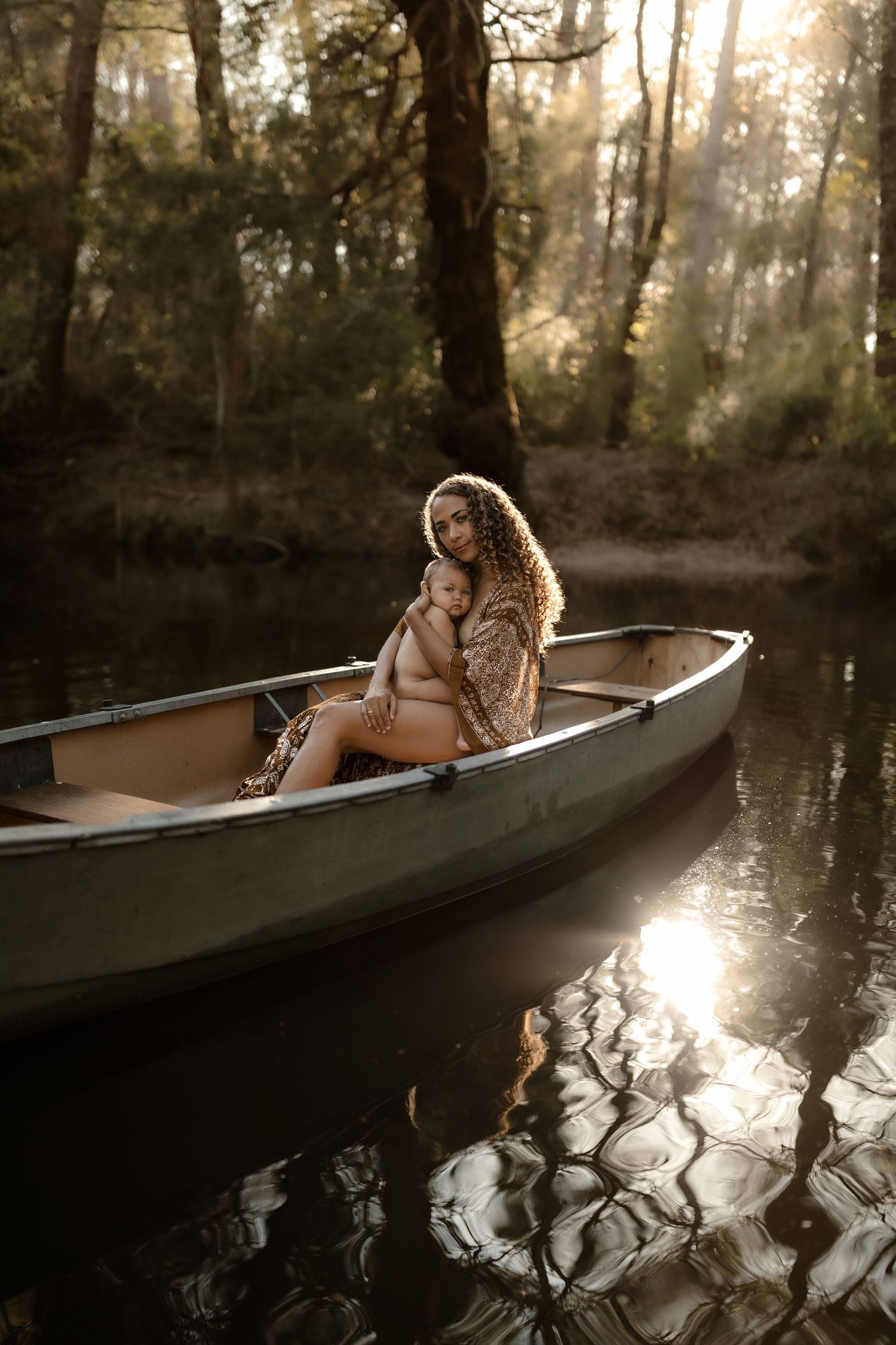 A curly-haired mother holds her baby while sitting in a canoe on a serene forest river at golden hour.