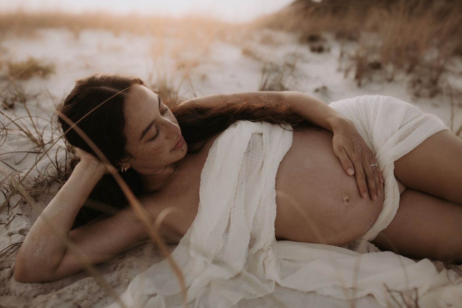 Pregnant woman in white fabric lying on sandy beach in golden sunset light, cradling her baby bump.