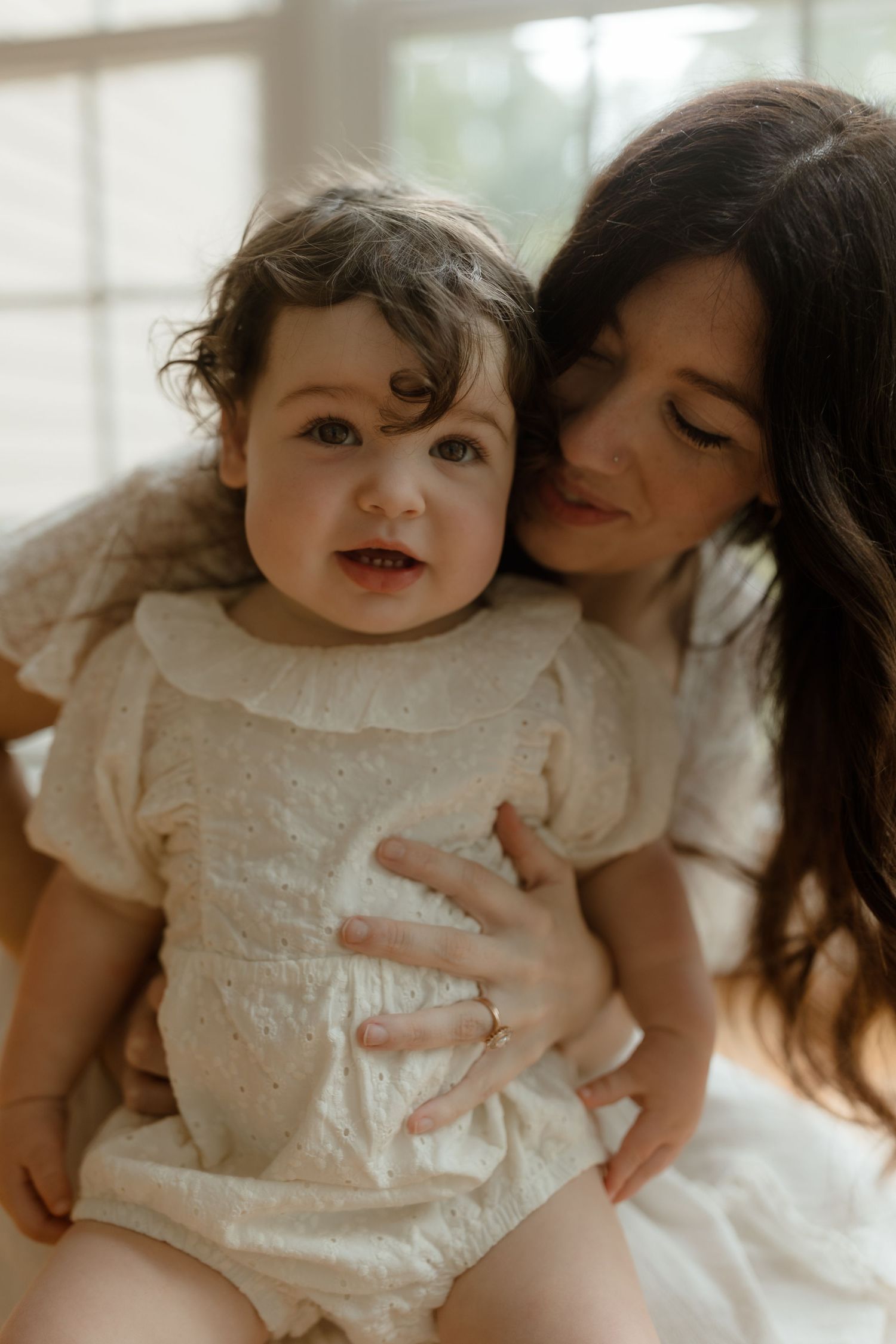 A mother in a white dress tenderly holds her baby girl in a cream eyelet romper near a bright window.