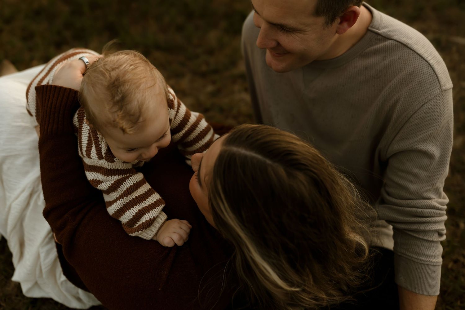 A smiling couple holds their baby outdoors in a warm family portrait session.