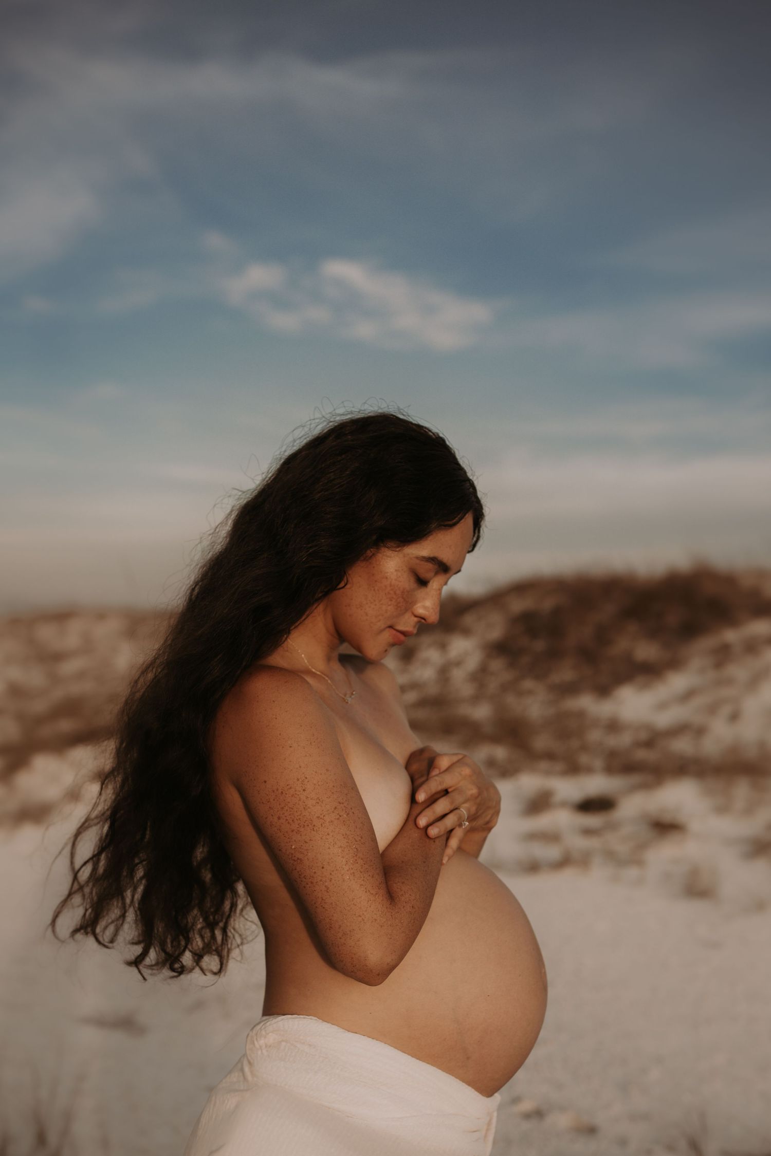 Pregnant woman with long dark hair holds her belly outdoors on sandy dunes under a blue sky.