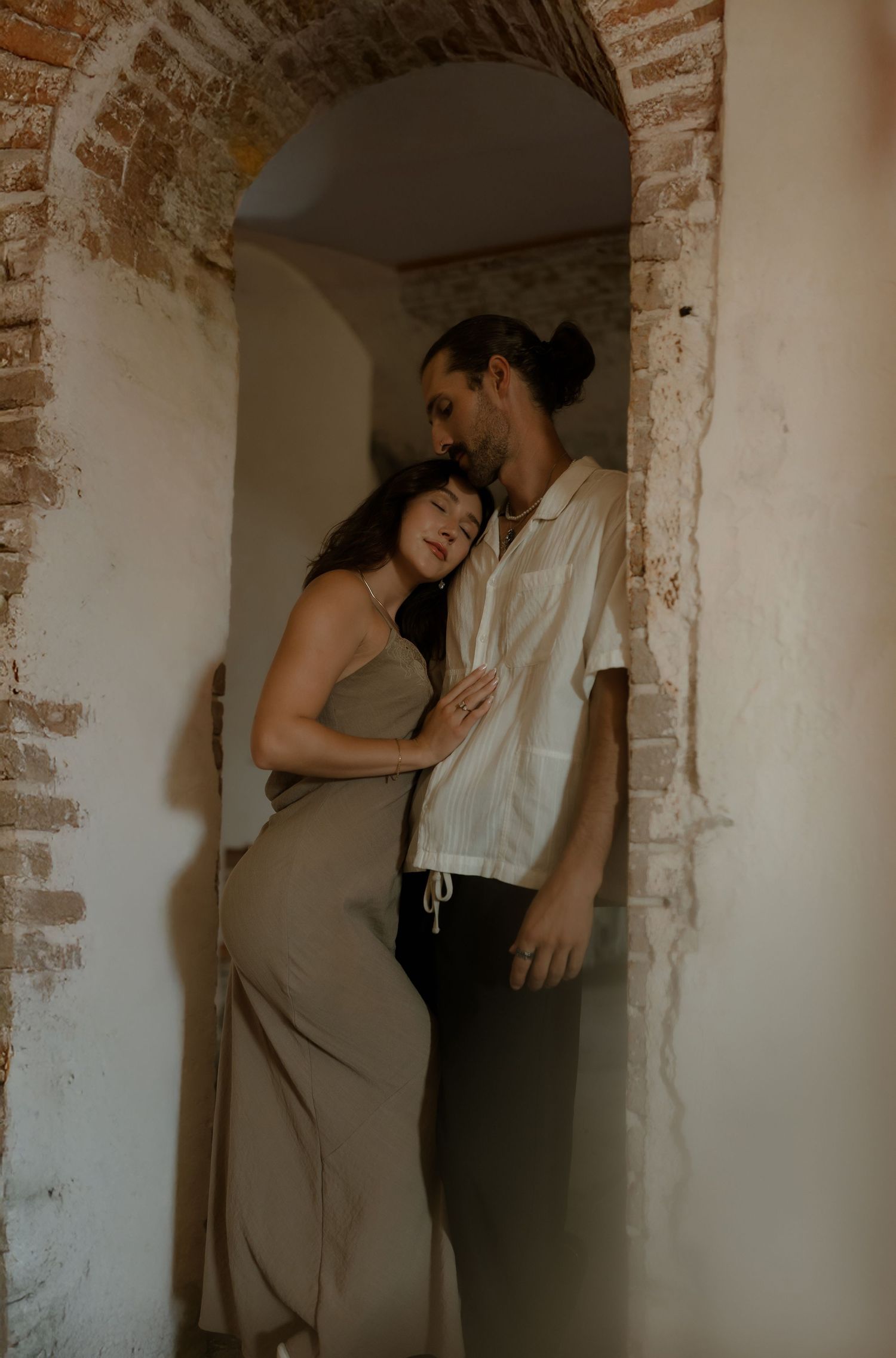 A couple shares an intimate moment in a rustic brick archway, the woman in a tan dress leaning into a man in a white shirt.