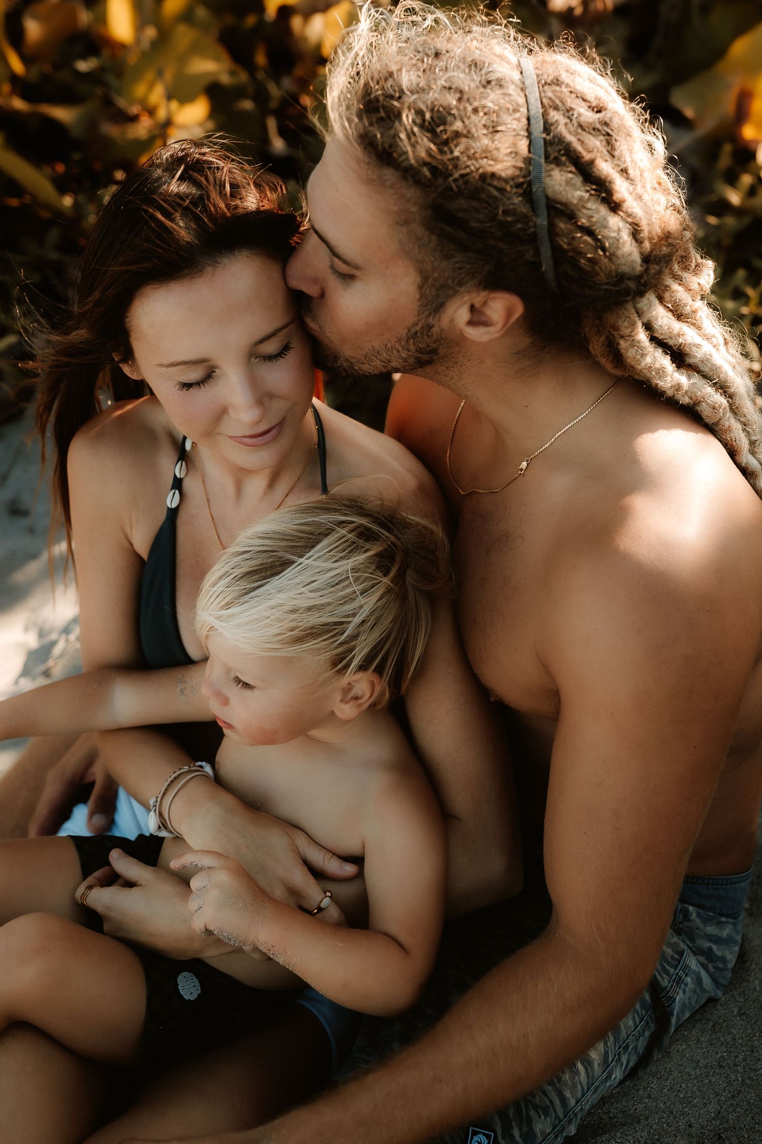 Family portrait of parents and toddler sitting together outdoors, man with dreadlocks kissing woman while child rests between them.