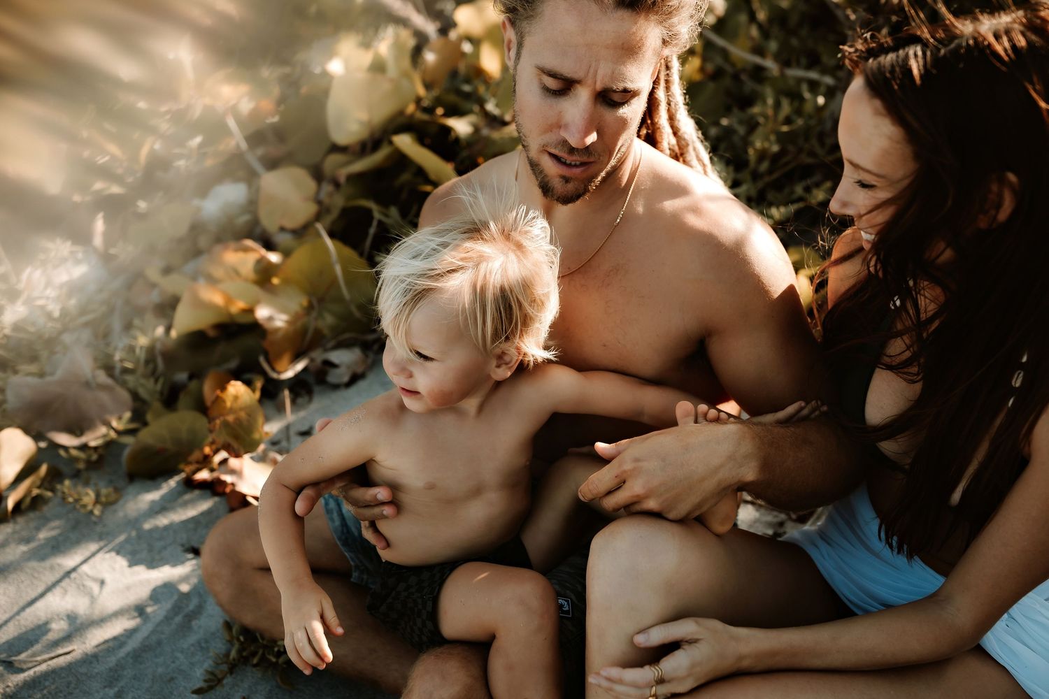 A shirtless father and mother sit outdoors with their young toddler, sharing a warm family moment in golden sunlight.