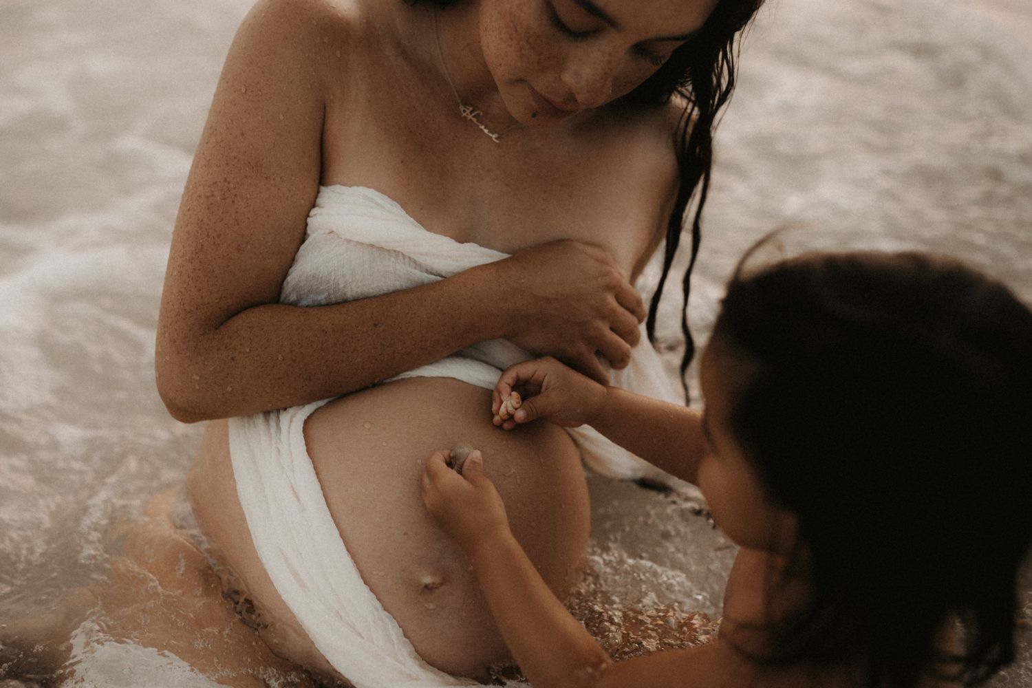 Pregnant mother in white wrap sits in shallow water as toddler touches her baby bump.