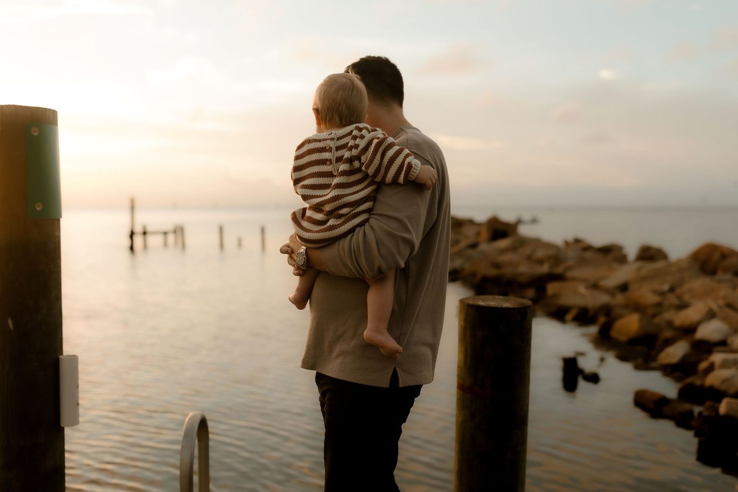 Father holding baby on rocky waterfront pier at sunset, sharing a tender moment by the calm sea.