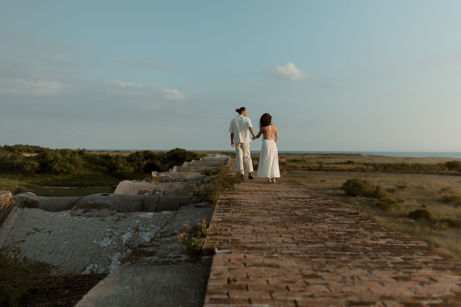 Couple in white walking hand in hand on a brick path toward the ocean at sunset.