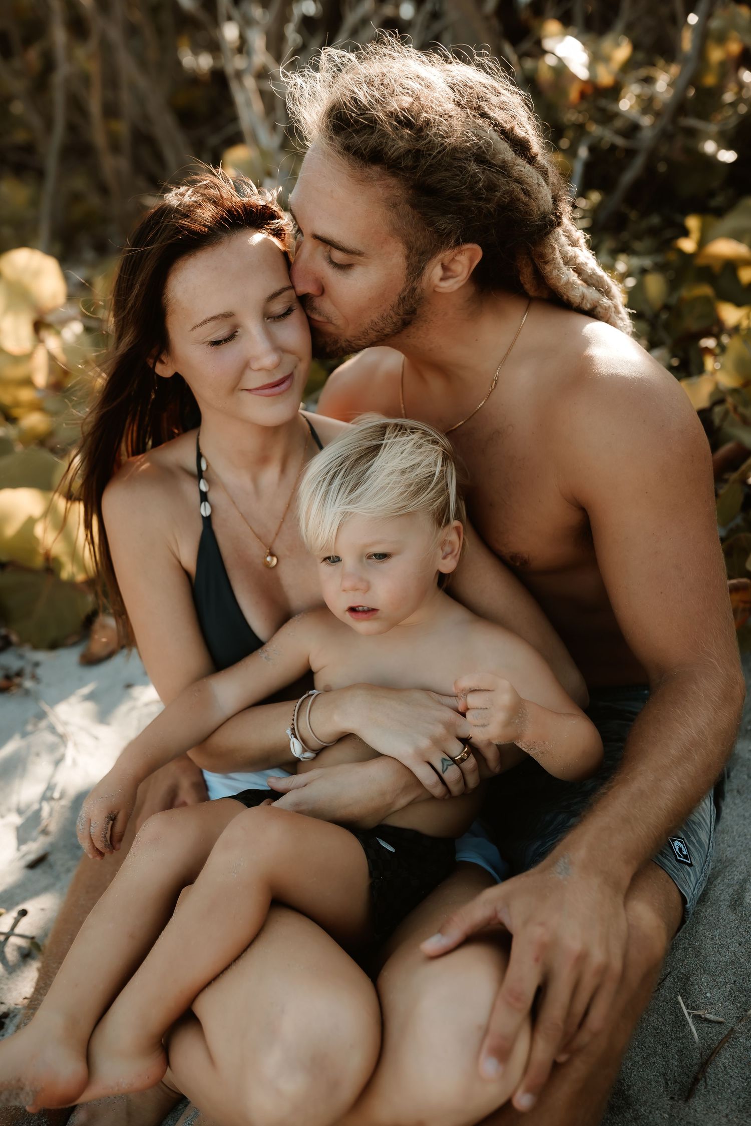 A happy family with a young toddler sitting together on a sandy beach surrounded by tropical foliage.