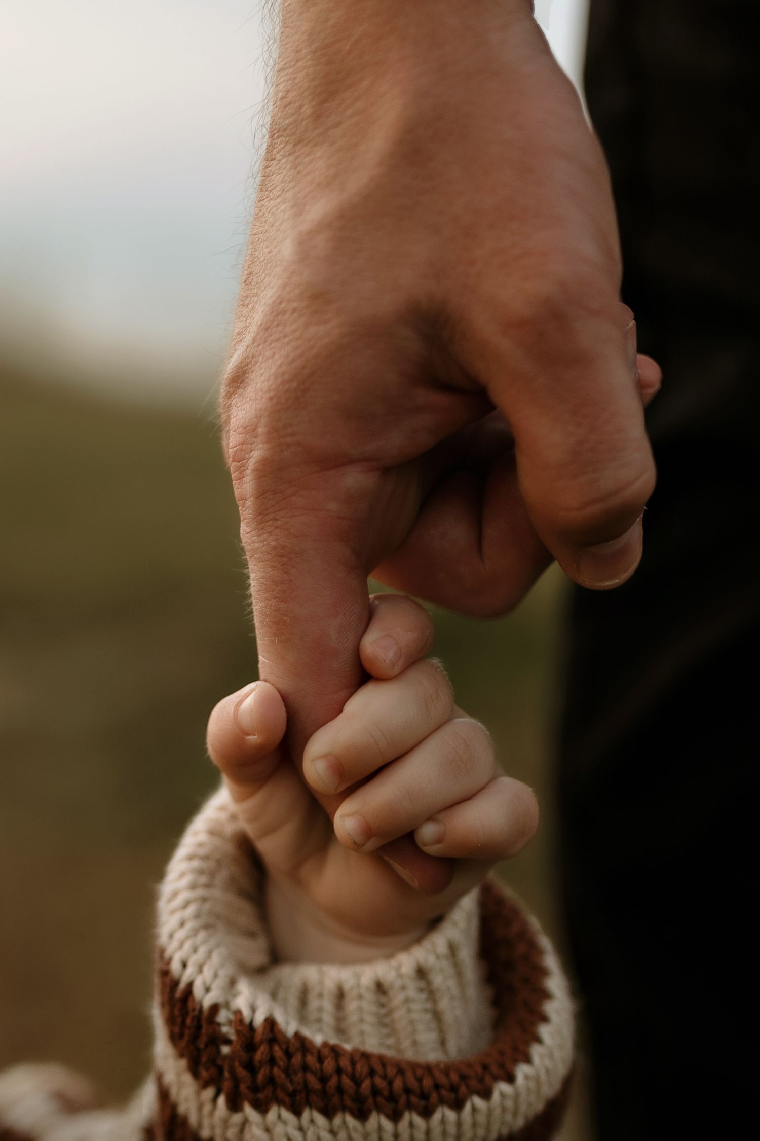 A parent's hand gently holds a baby's tiny fist, wearing a knit sweater, in a tender close-up moment.