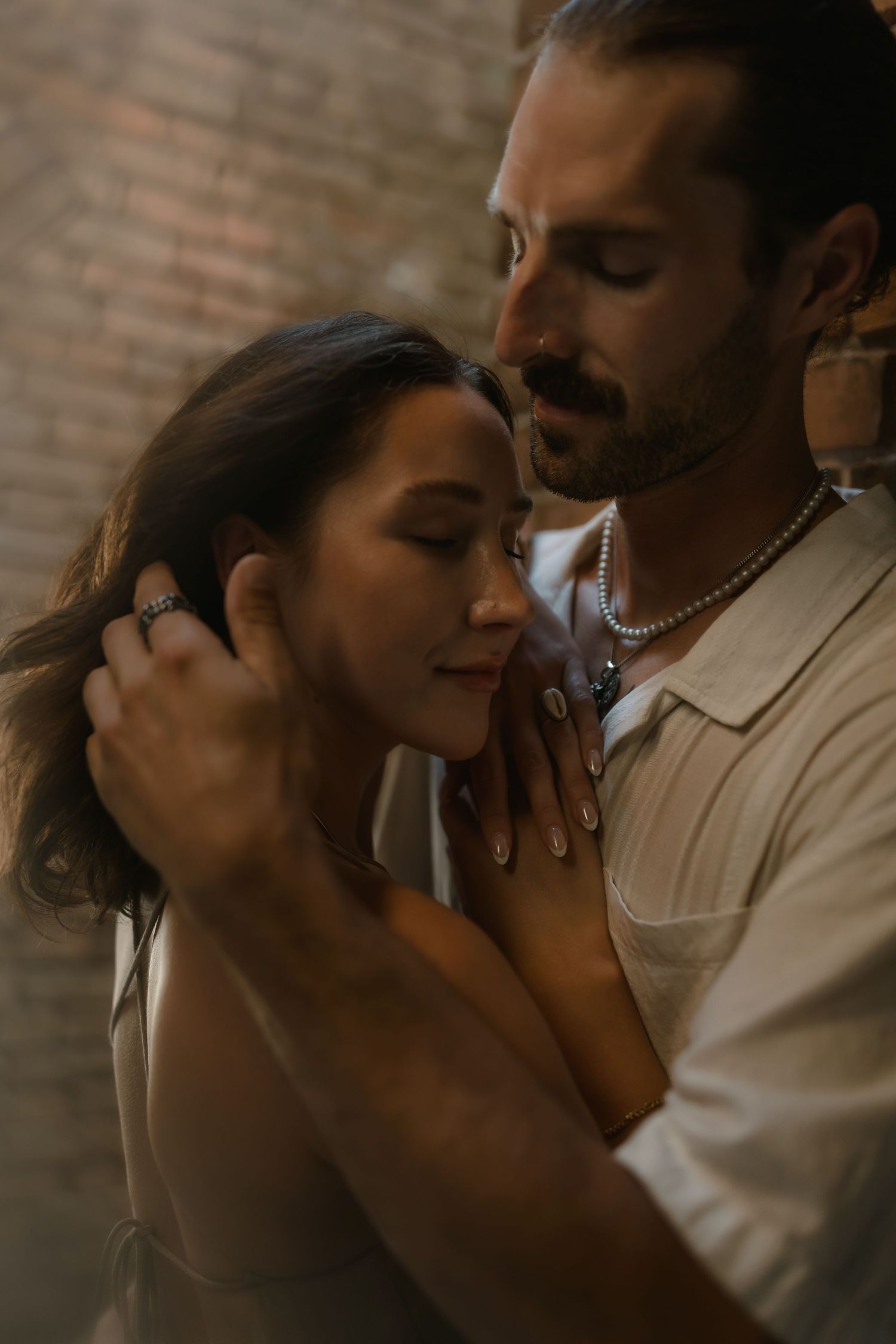 A couple shares an intimate embrace, the man gently cradling the woman's face as both close their eyes near a brick wall.