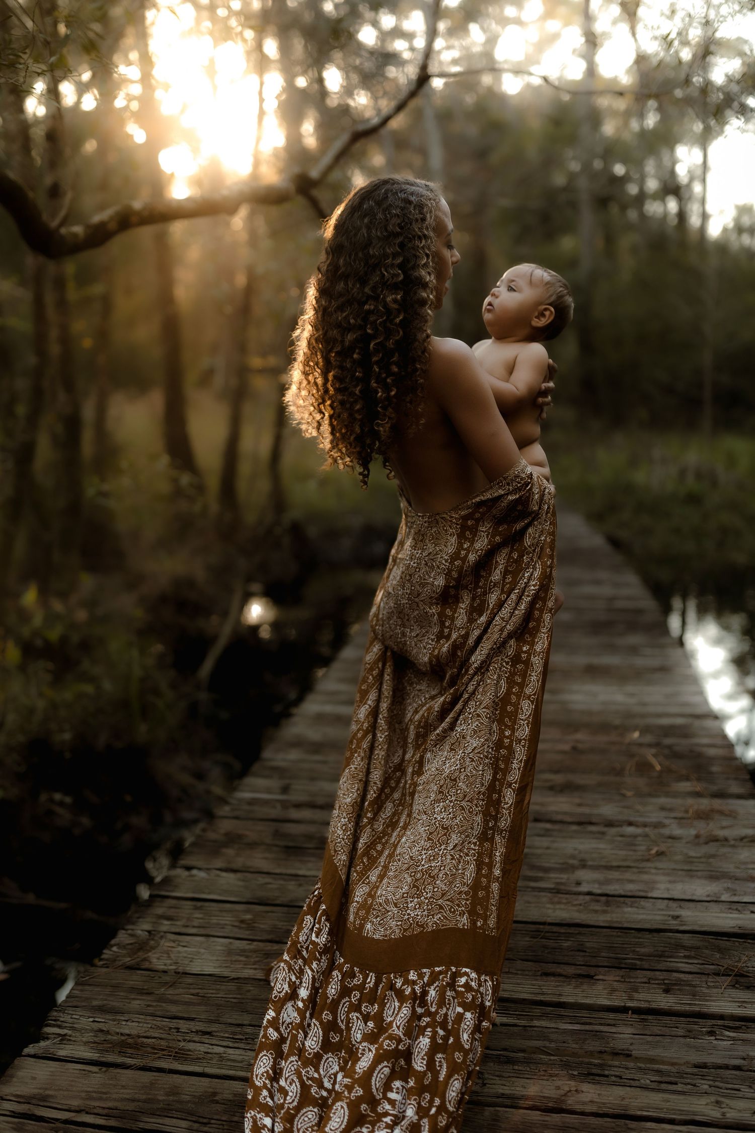 Mother holding newborn baby on wooden bridge at golden sunset in forest, wearing floral wrap dress with curly hair.