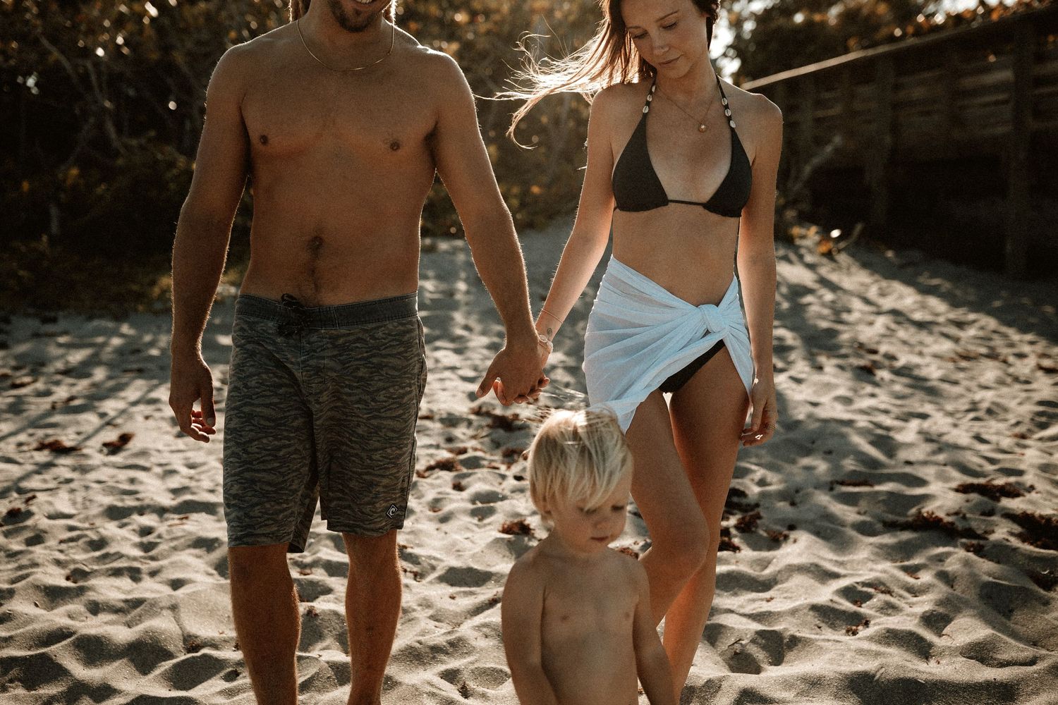 Family of three walking on sandy beach, parents in swimwear holding toddlers hands in golden sunlight.
