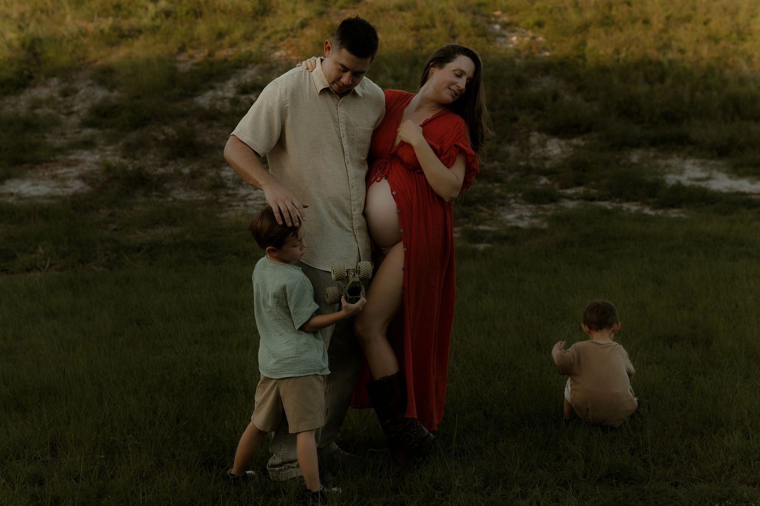 Pregnant woman in red dress poses outdoors with partner and two young children during golden hour maternity photo session.