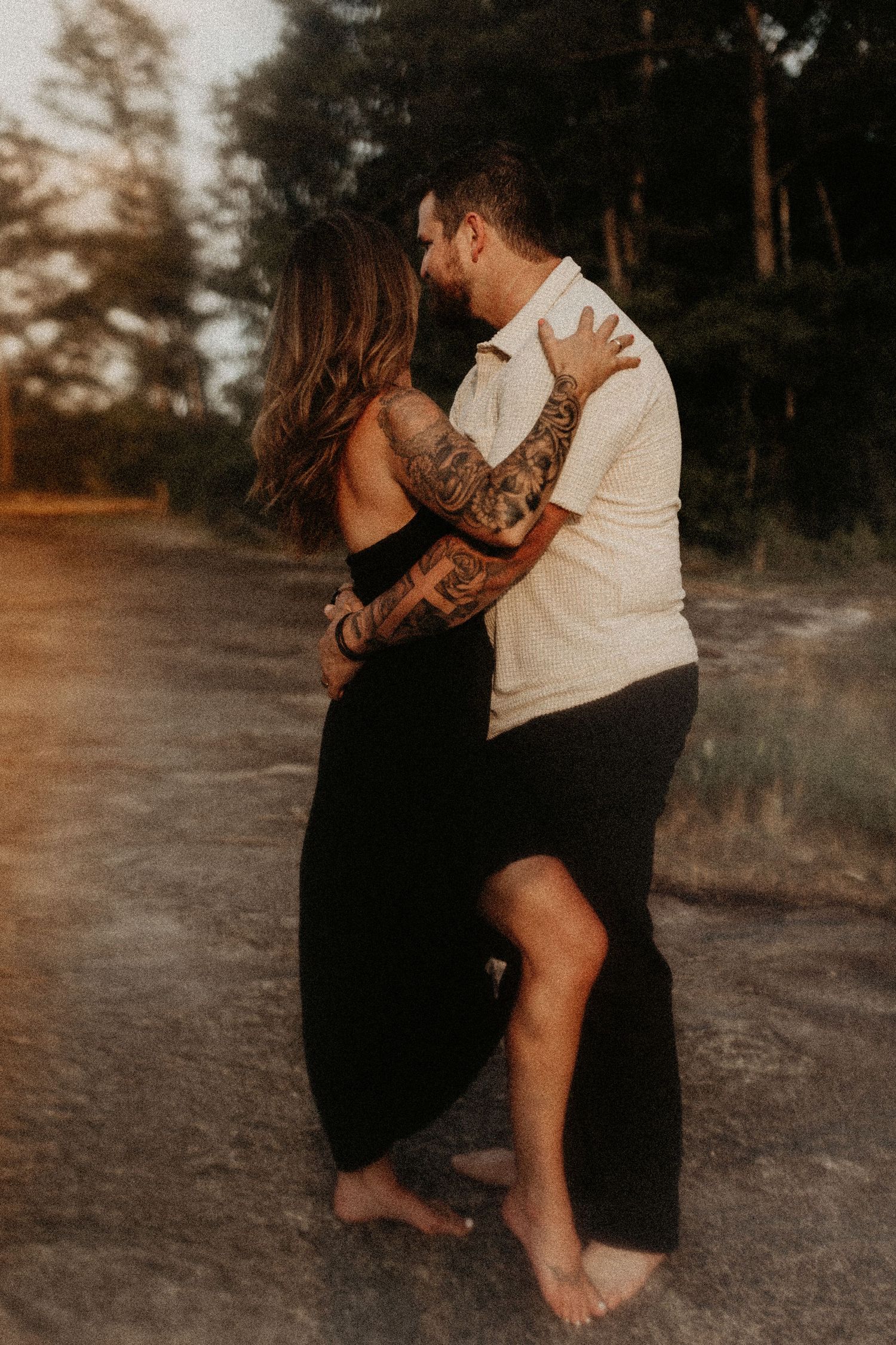 Tattooed couple sharing a romantic kiss while embracing barefoot on a dirt path surrounded by trees.