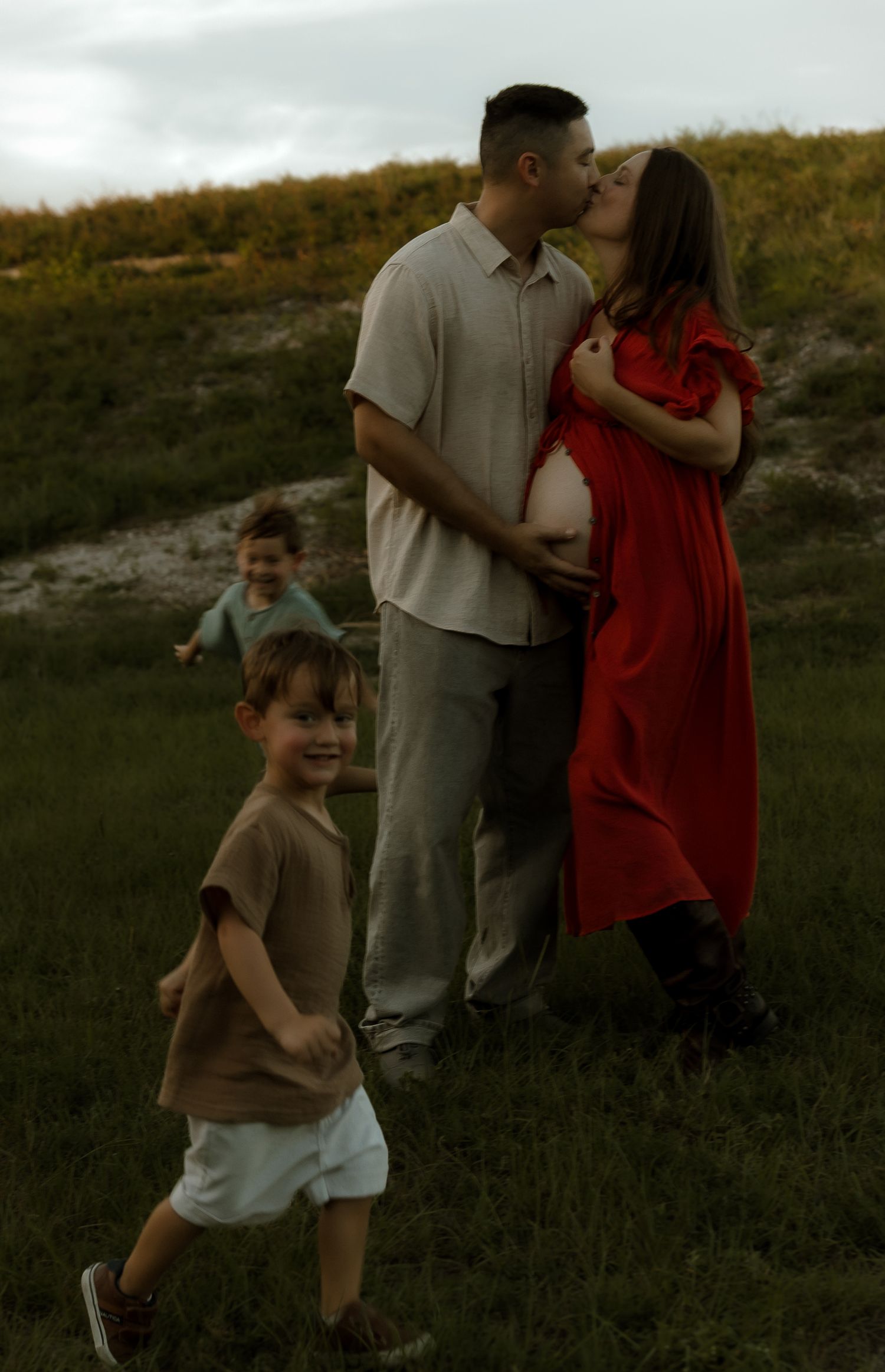 Pregnant woman in red dress kissing partner while two young children play nearby in grassy field.