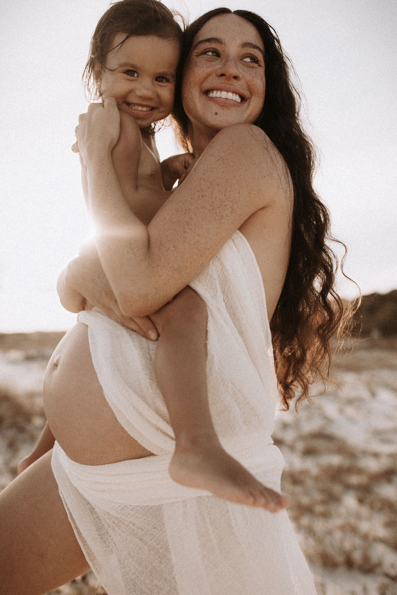 Pregnant woman with freckles holding toddler on beach, both smiling in white wrap.