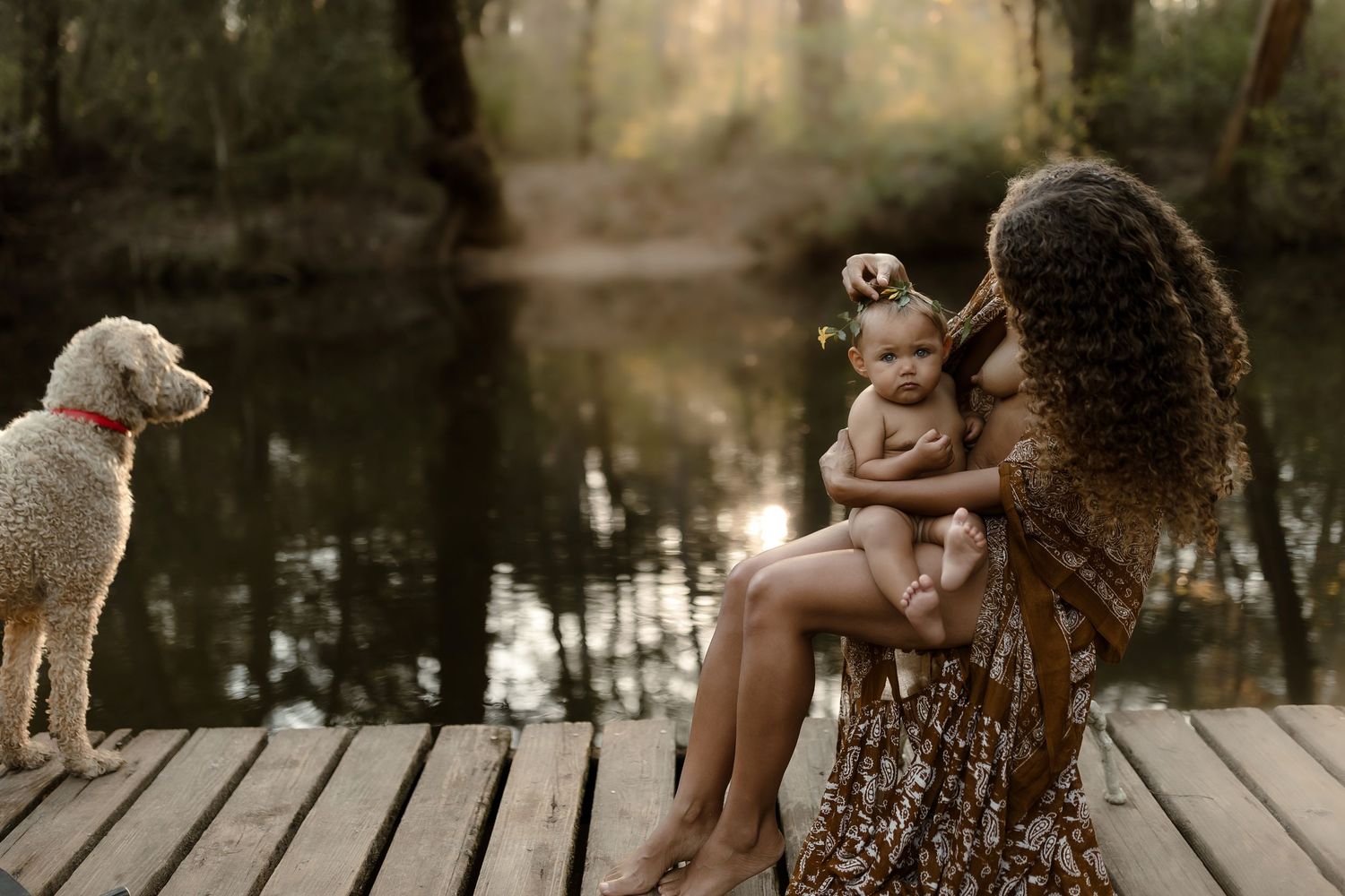 Mother holding baby on wooden dock near water with white poodle nearby in natural forest setting.