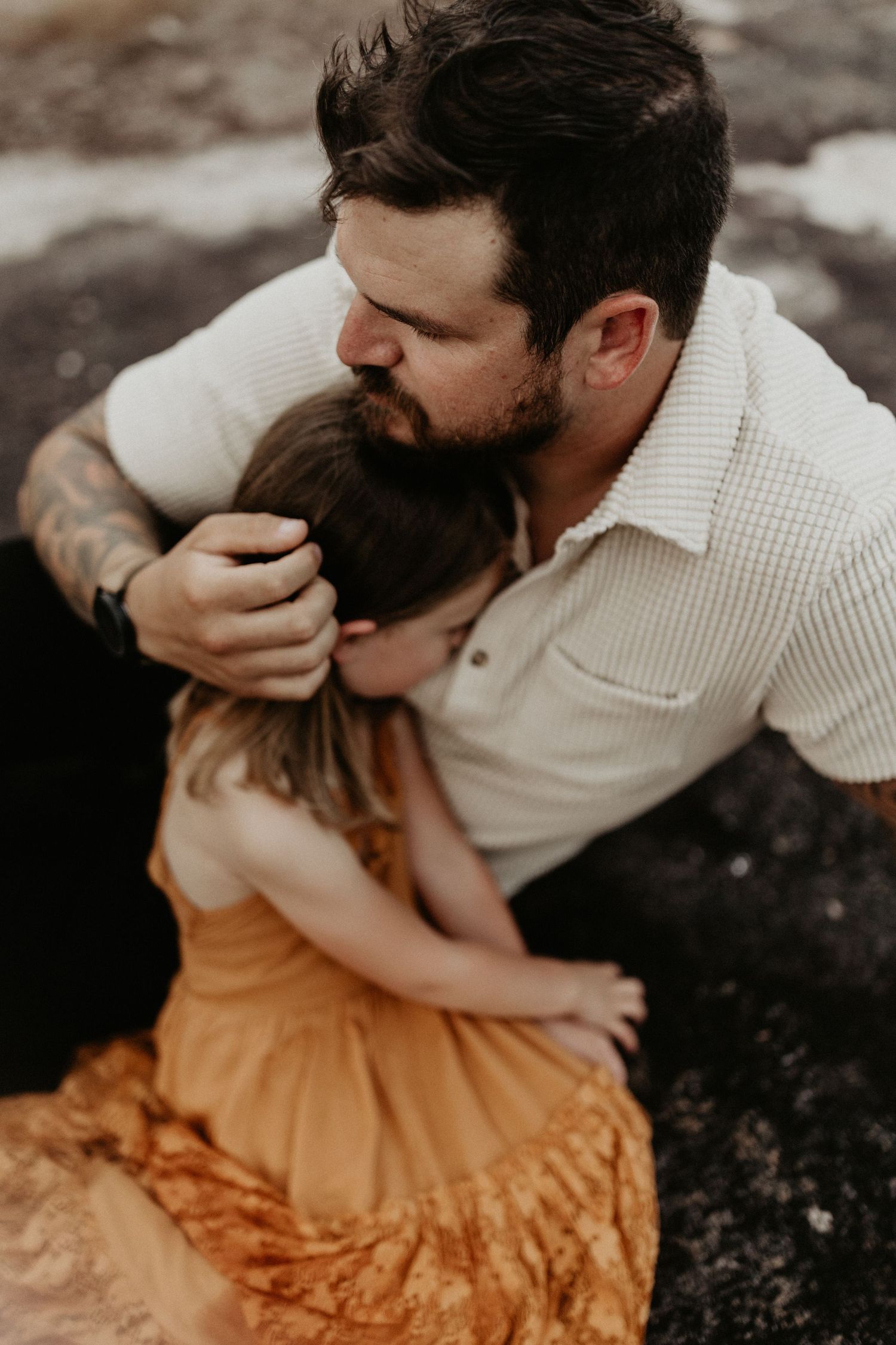 A bearded man with tattoos tenderly kisses a woman in an orange dress during a romantic outdoor couples session.