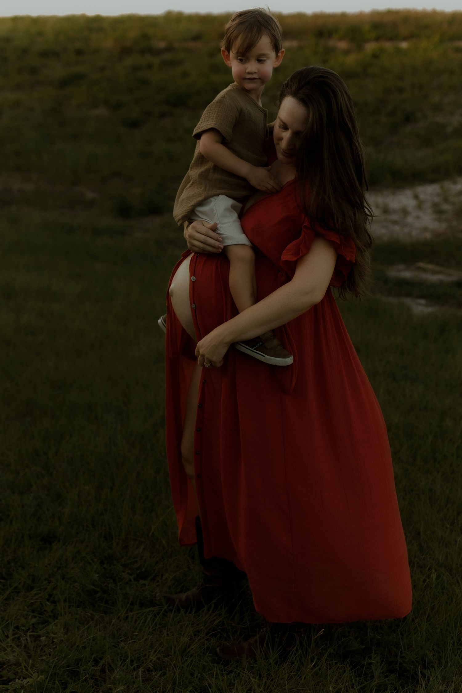 Pregnant woman in red dress holds toddler boy outdoors on green grass at golden hour.