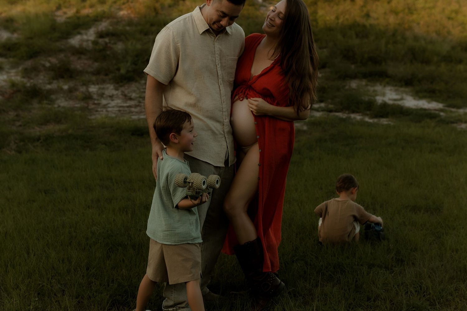 Pregnant woman in red dress poses with partner and young son in a grassy field during golden hour maternity photo session.