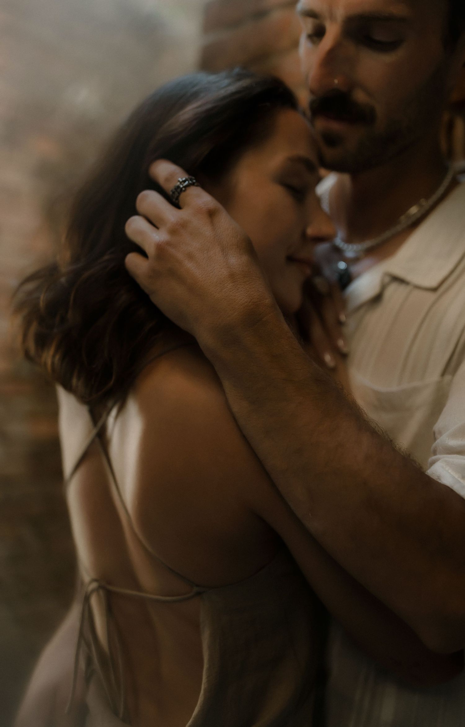 A couple shares an intimate embrace, the man tenderly holding the woman close with eyes closed in a warm, romantic moment.