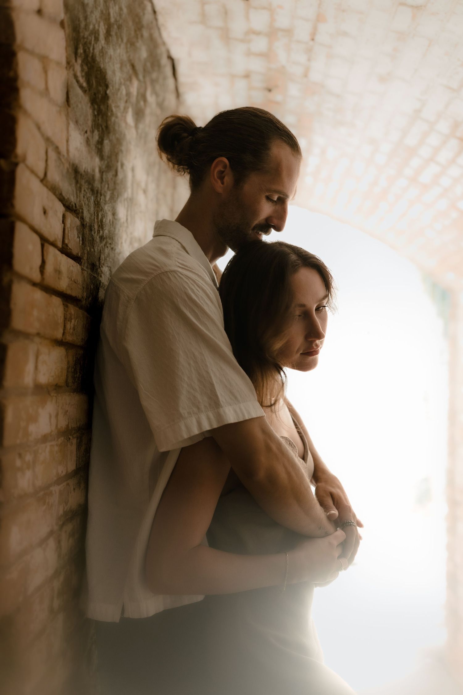 A couple embraces tenderly in a brick archway, soft light illuminating their peaceful moment together.