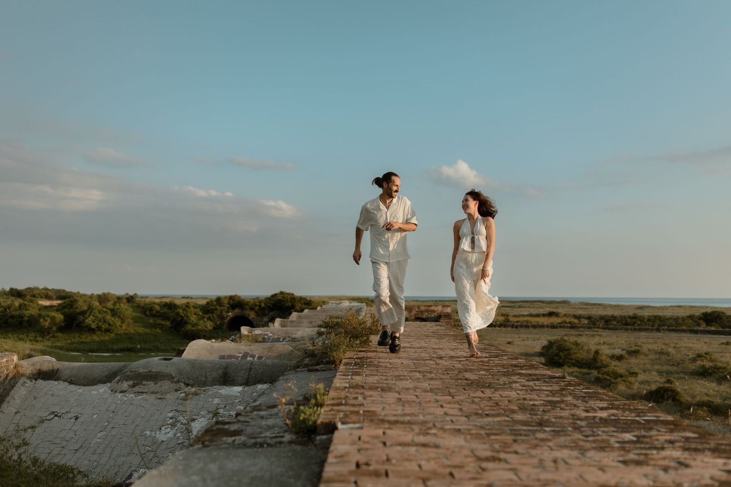 A couple in white attire joyfully runs along a stone path with a scenic coastal landscape under a clear blue sky.