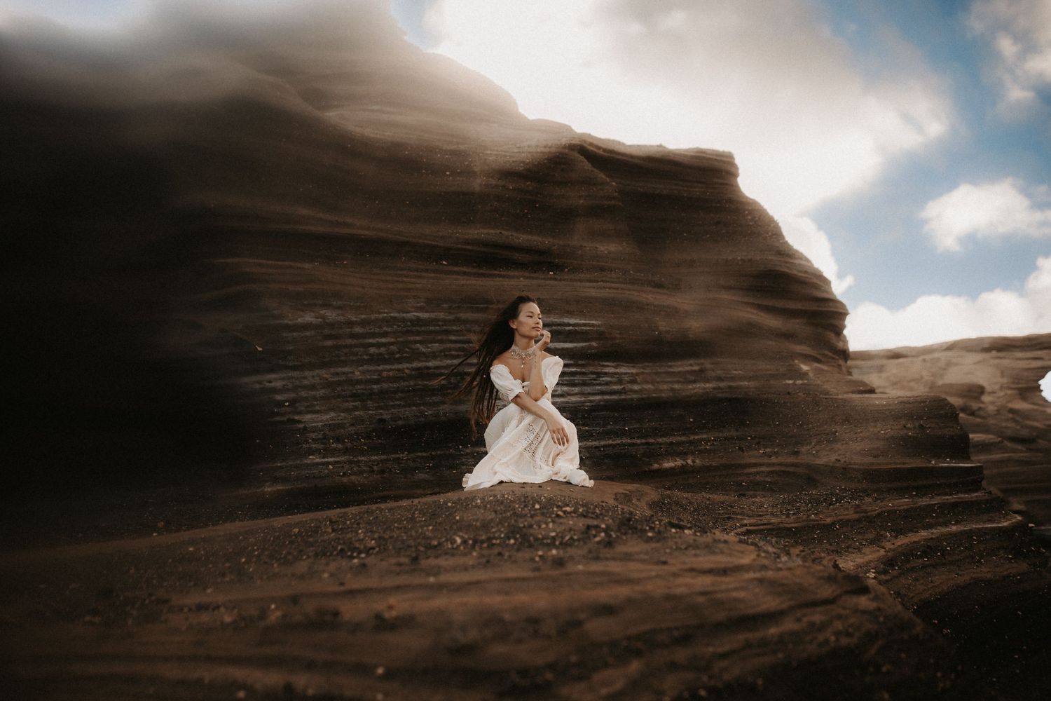 A woman in a white dress stands against dramatic layered rock formations under a cloudy sky.