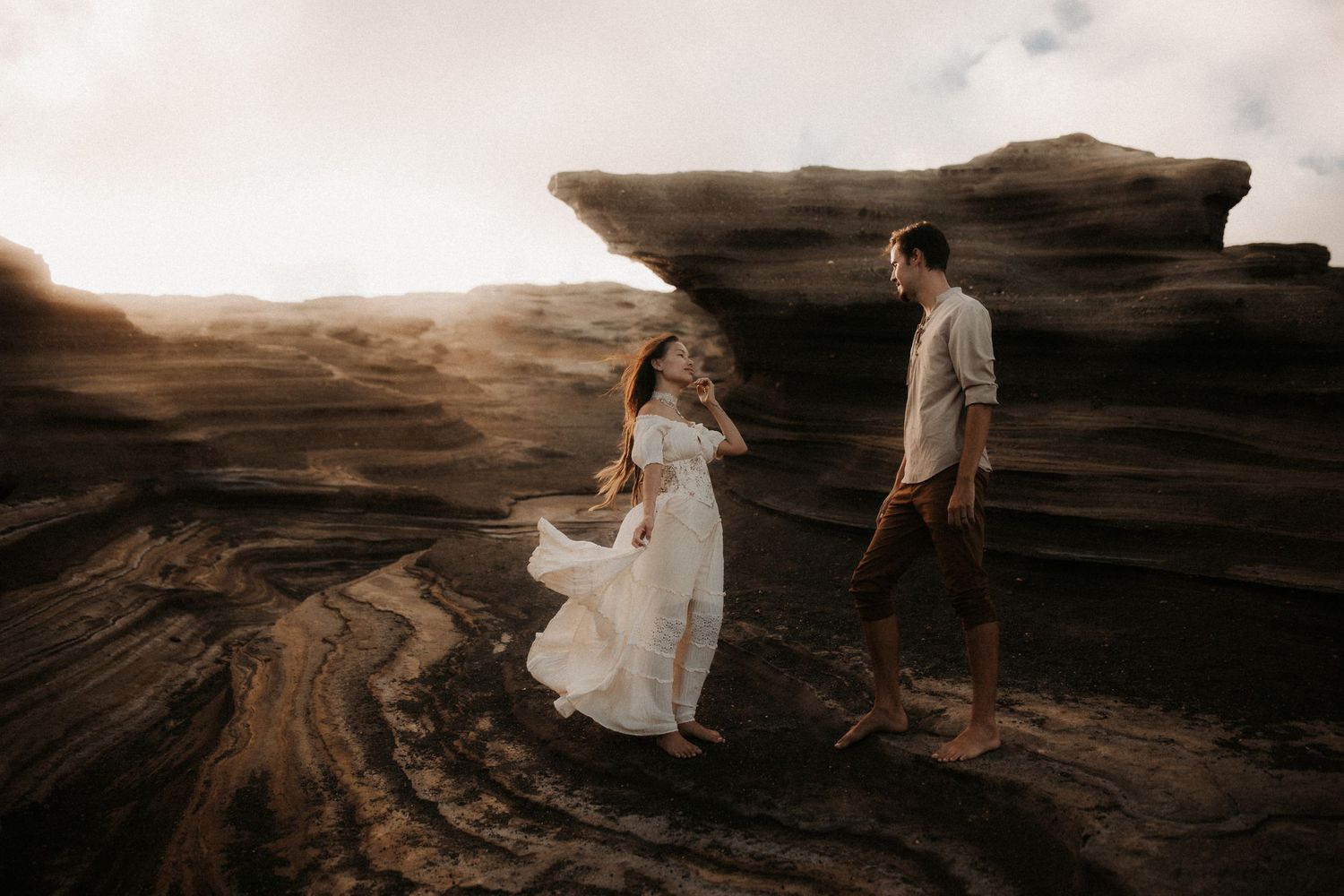 Couple walking on dramatic volcanic rock formations at sunset, woman in flowing white dress, man in casual attire.
