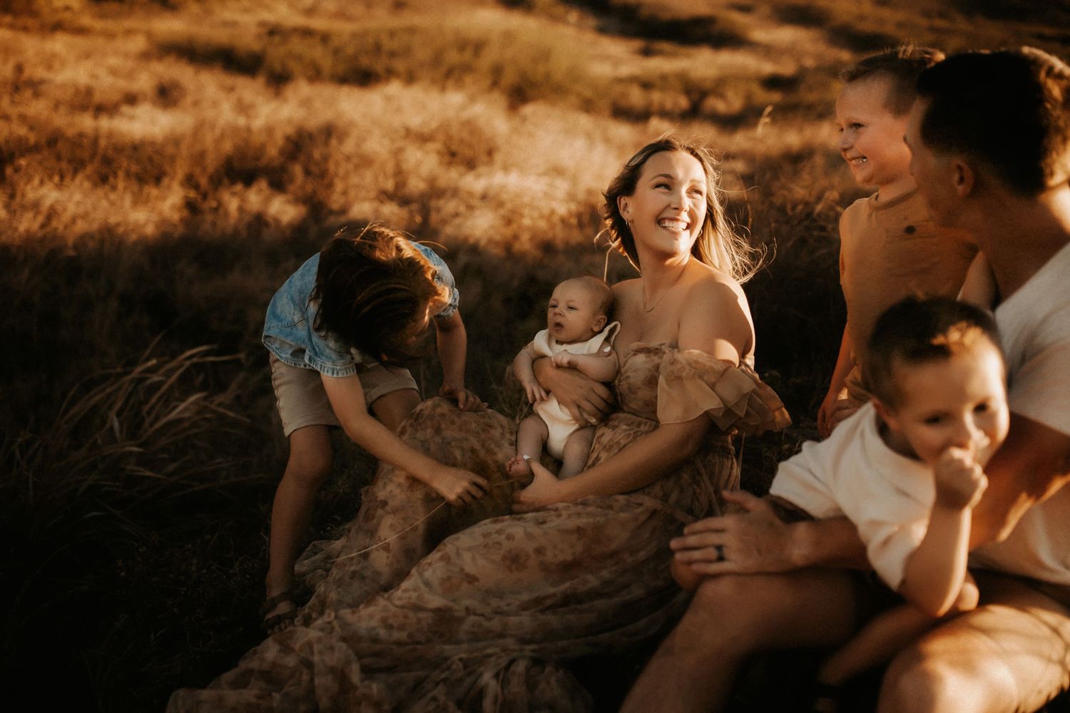 Mother laughing joyfully with children and baby in a golden sunlit field.