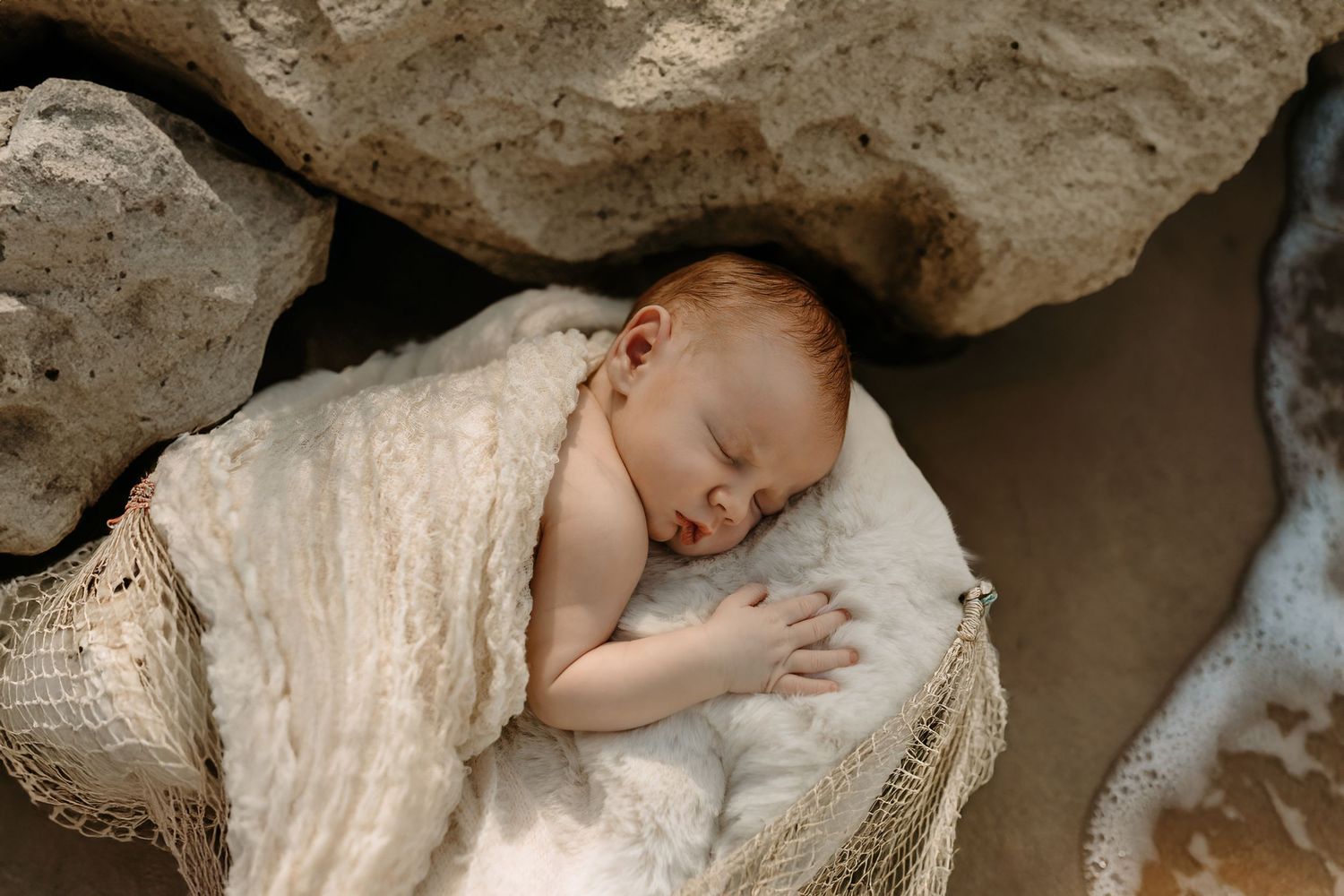 Sleeping newborn baby wrapped in cream blanket nestled between rocks outdoors.