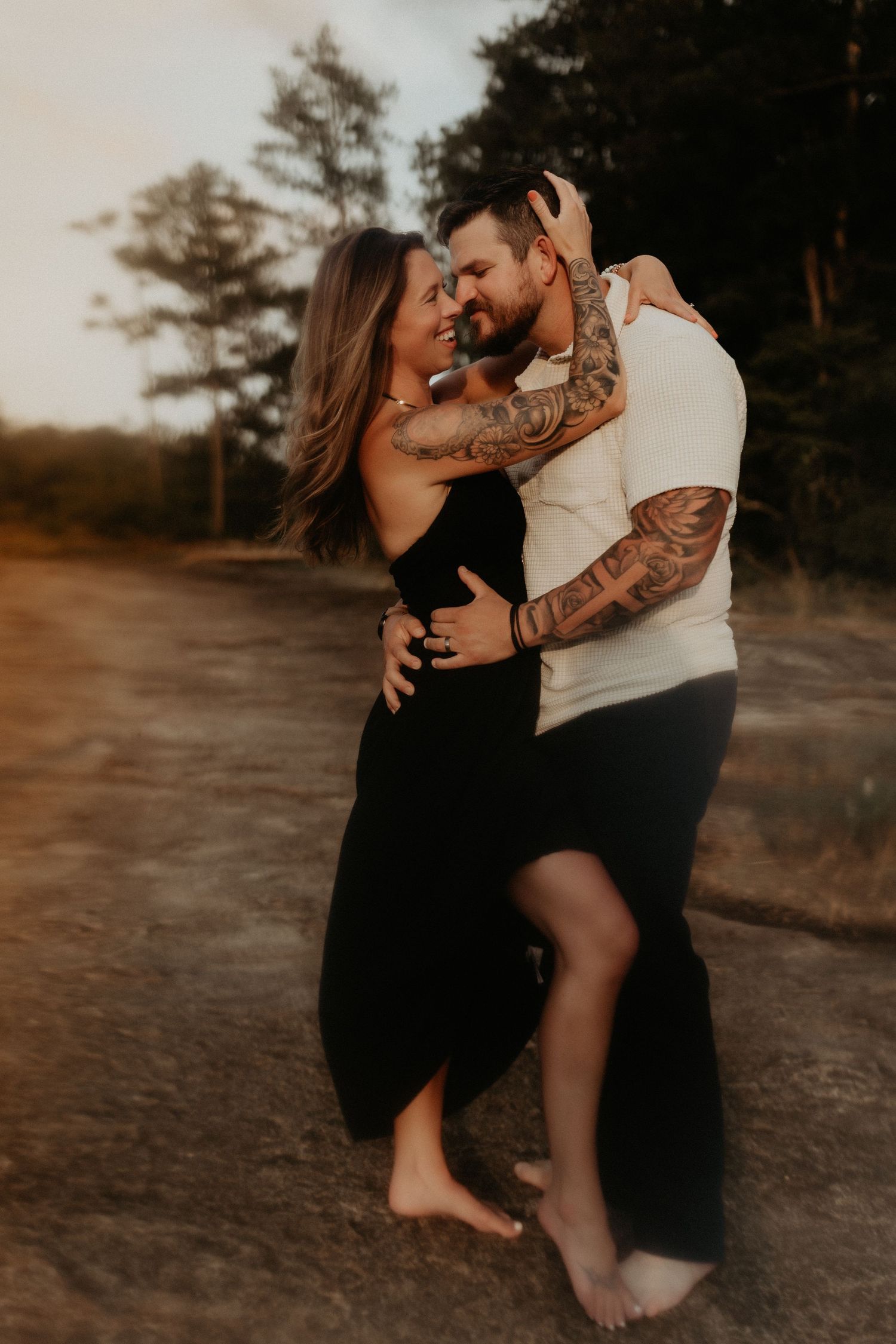 Couple dancing barefoot outdoors at golden hour, woman in black dress embraced by tattooed man in white shirt.