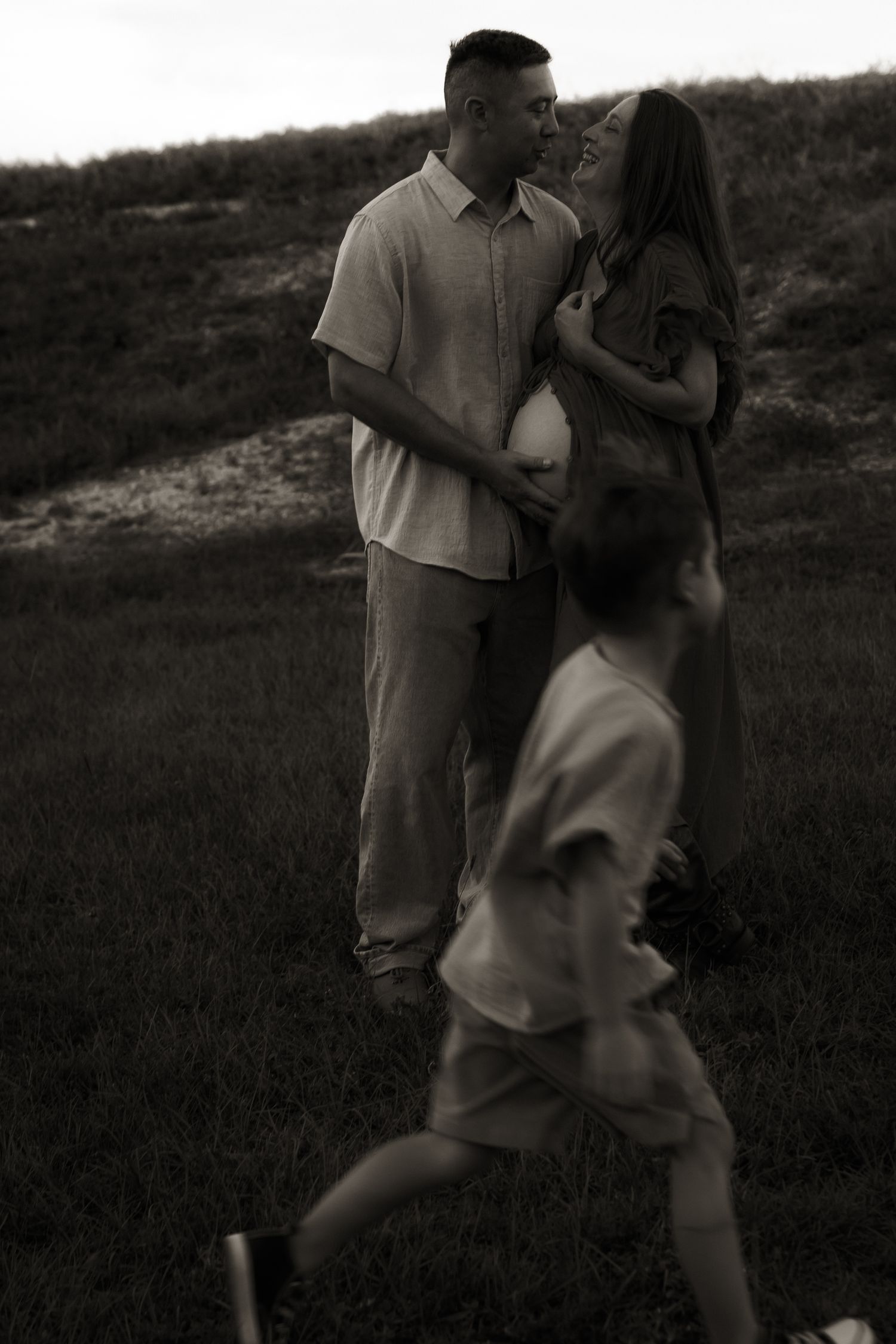 A couple shares a tender moment outdoors as a child runs past, with the woman visibly pregnant, in a moody black-and-white photo.