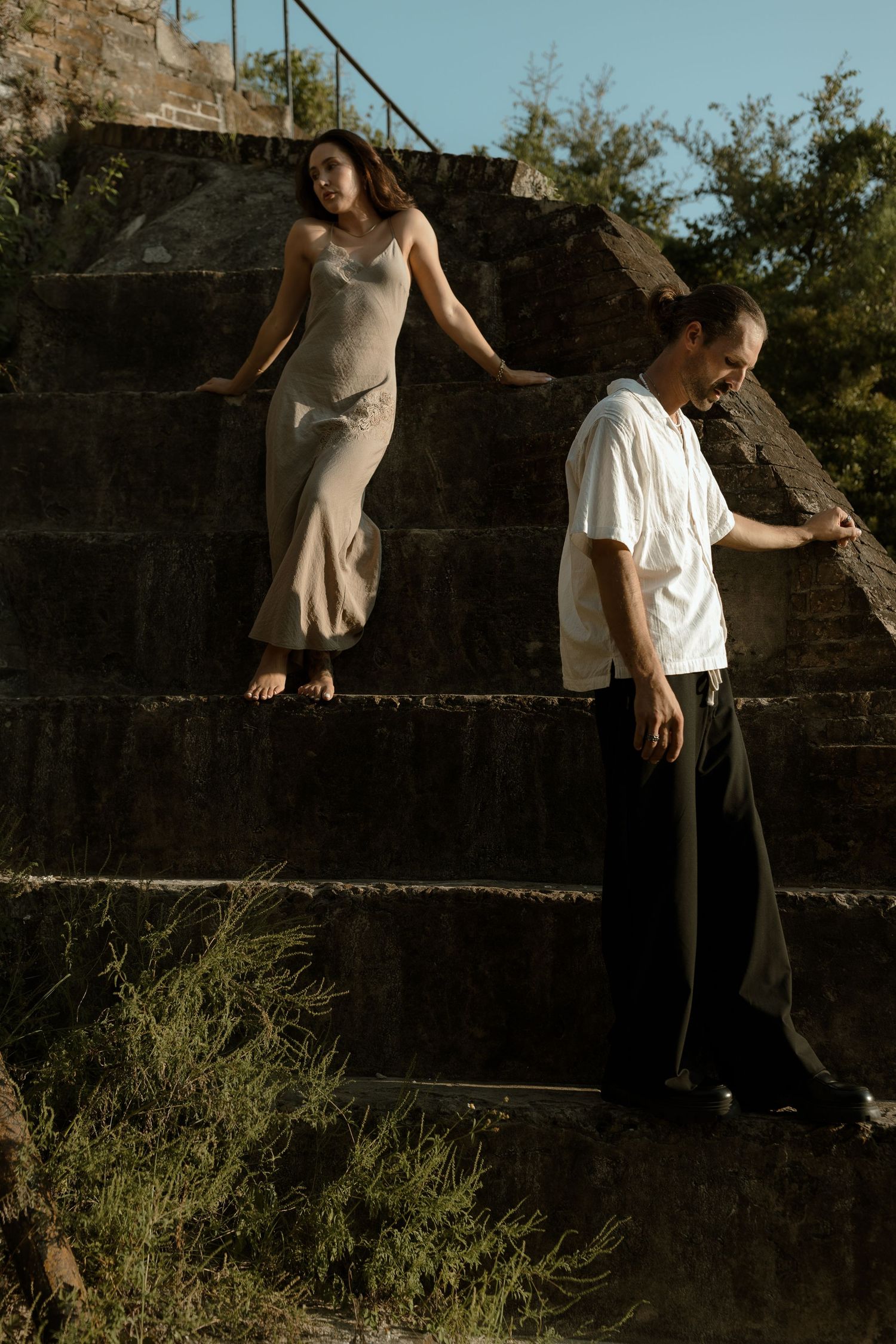 A woman in a slip dress stands on stone steps above a man in a white shirt and black pants outdoors.