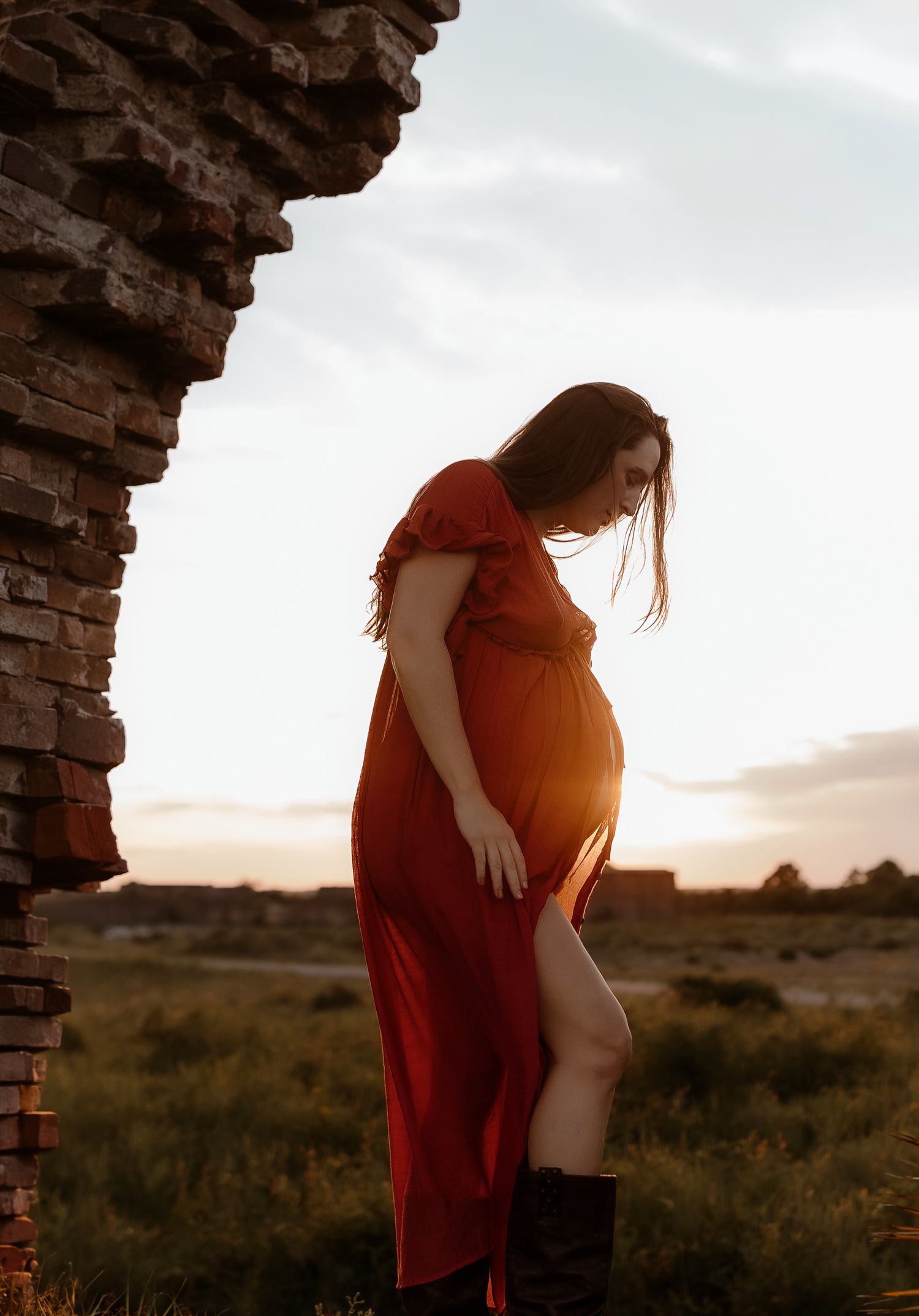 Pregnant woman in red dress and boots poses near brick arch at golden hour sunset in open field.