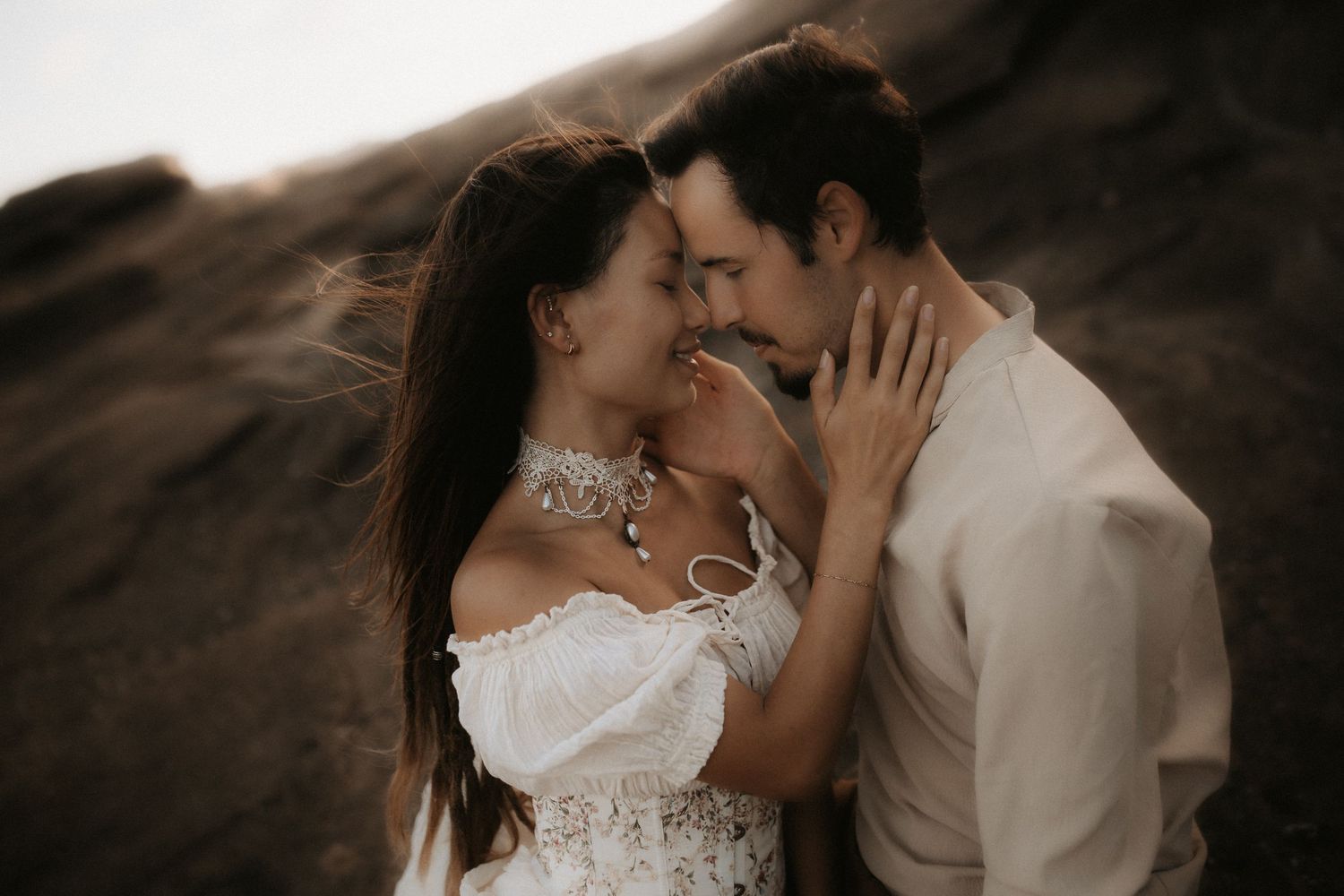 Romantic couple in white attire sharing an intimate moment against a rocky backdrop outdoors.