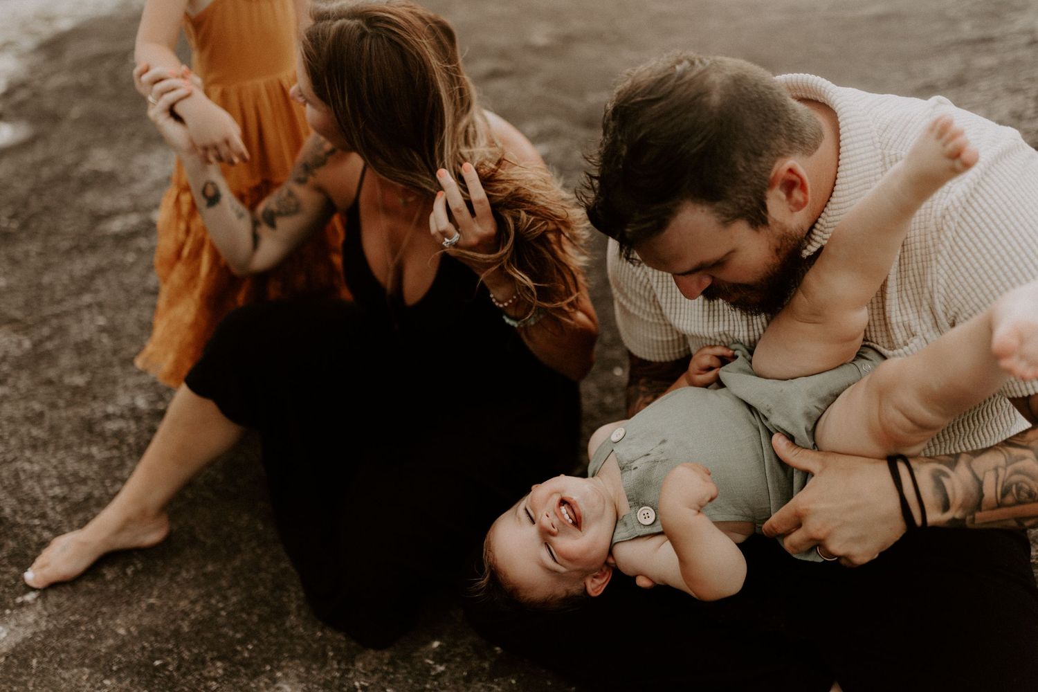 Playful family moment with tattooed parents laughing and tickling their baby on the ground outdoors.