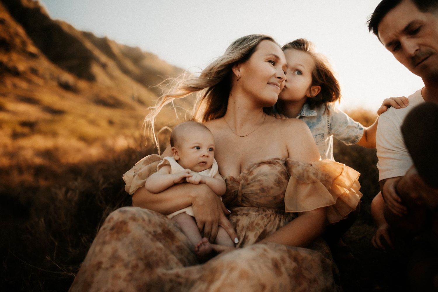 Family sitting outdoors at golden hour, mother holding baby and toddler, father beside them on a hillside.