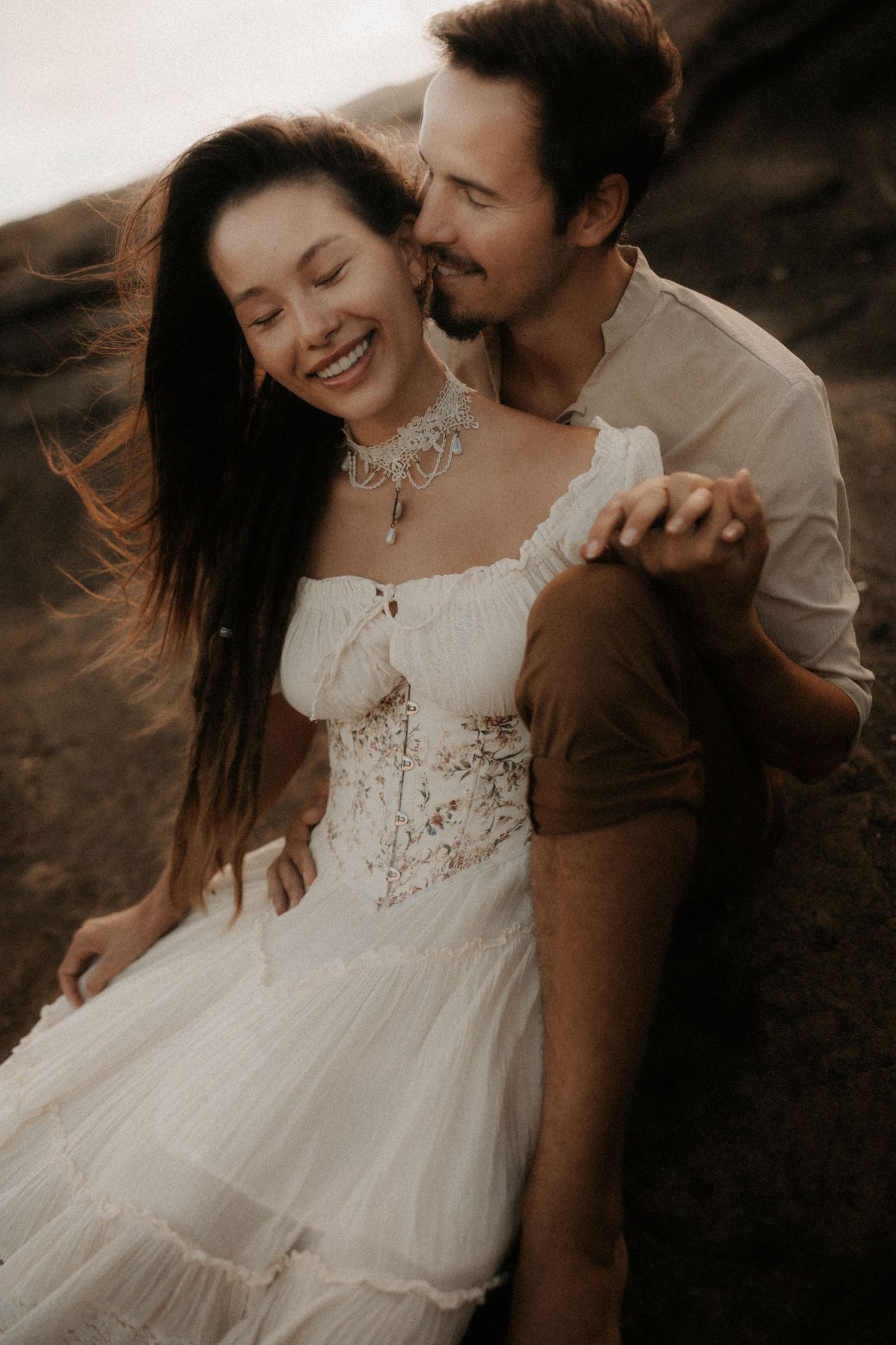 A couple in bohemian attire embraces outdoors, the woman in a white lace dress smiling joyfully as the man nuzzles her cheek.