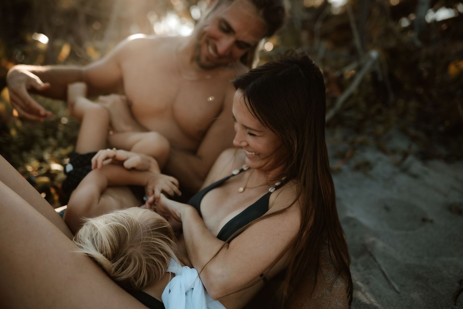 Happy family with young child laughing together on sandy beach in warm sunlight.