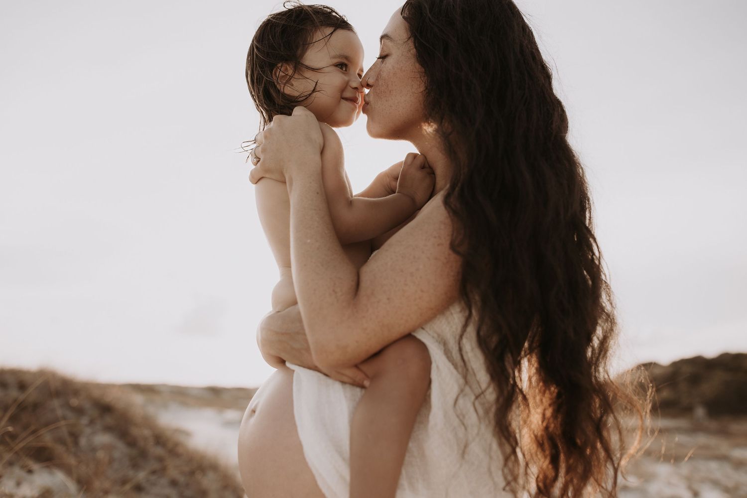 Pregnant mother with long dark hair kisses smiling toddler on sandy beach at sunset.