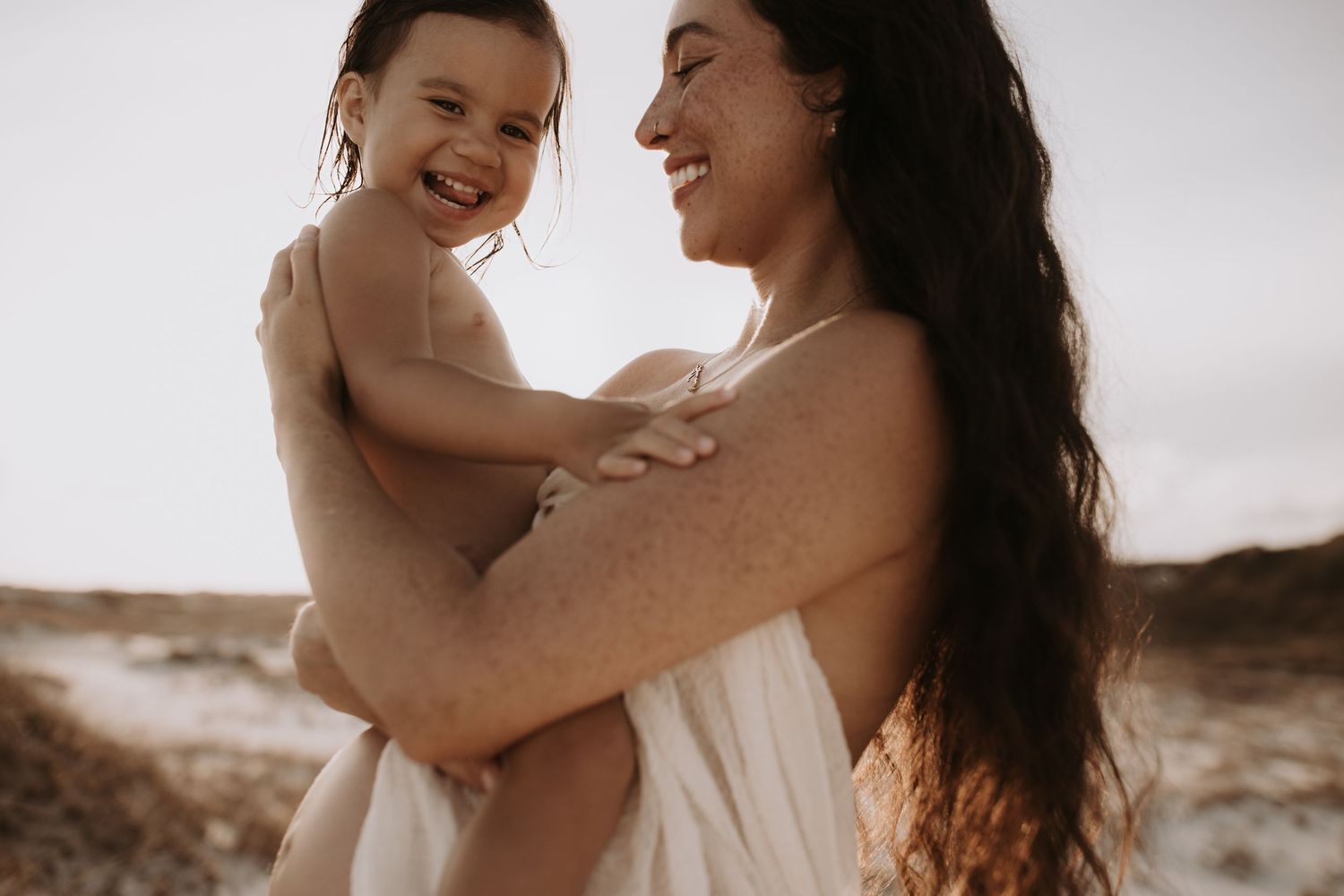 Laughing mother lifting smiling toddler girl at the beach in warm sunlight.