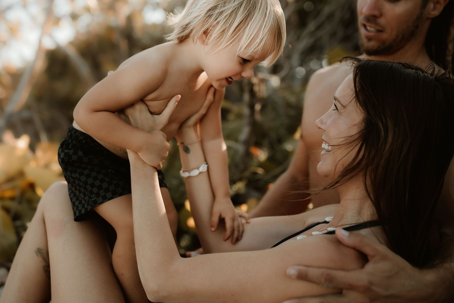 A young blonde toddler laughs while sitting on parents laps outdoors in warm golden light.