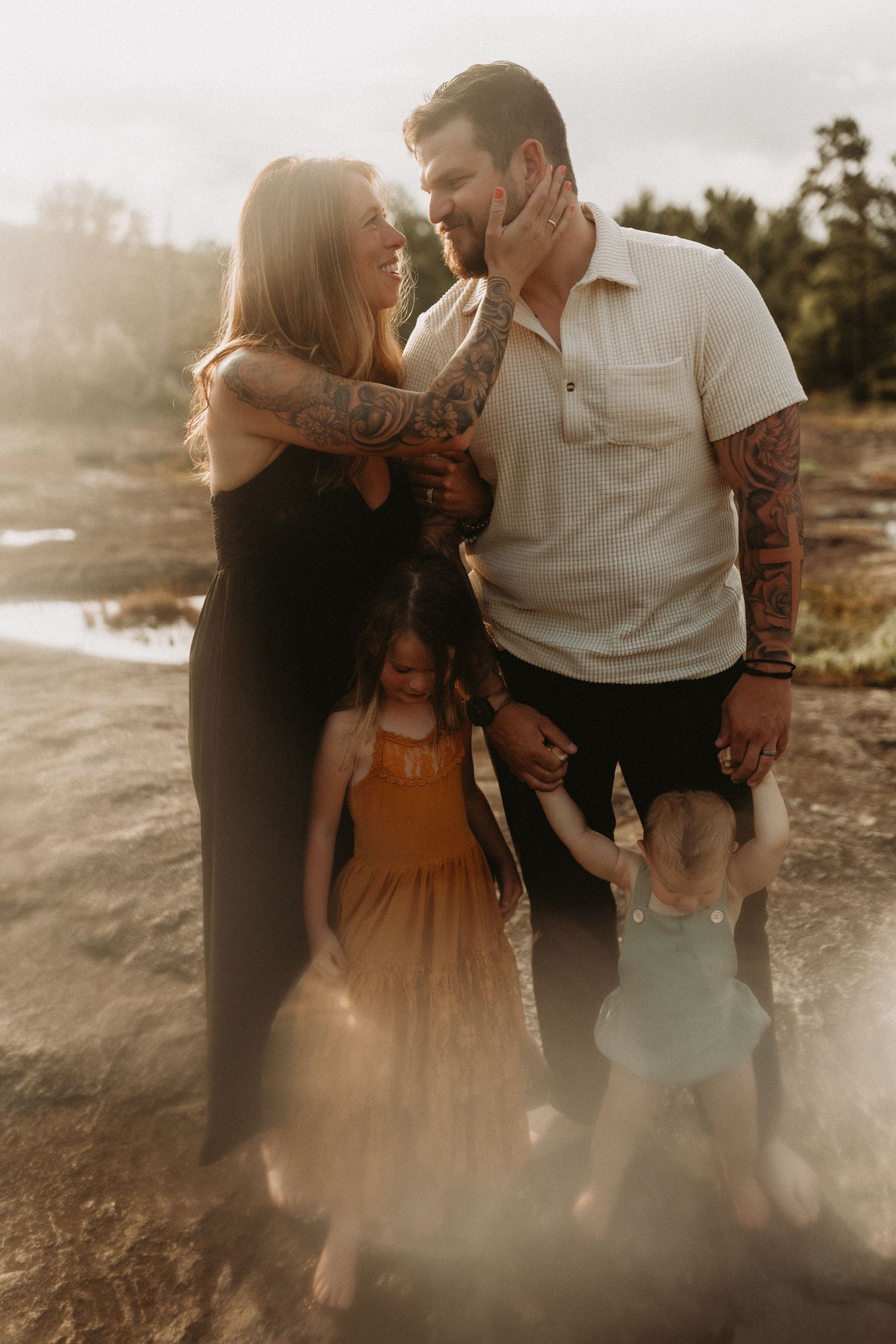 Tattooed couple smiling at each other outdoors at golden hour with two young daughters in boho dresses.