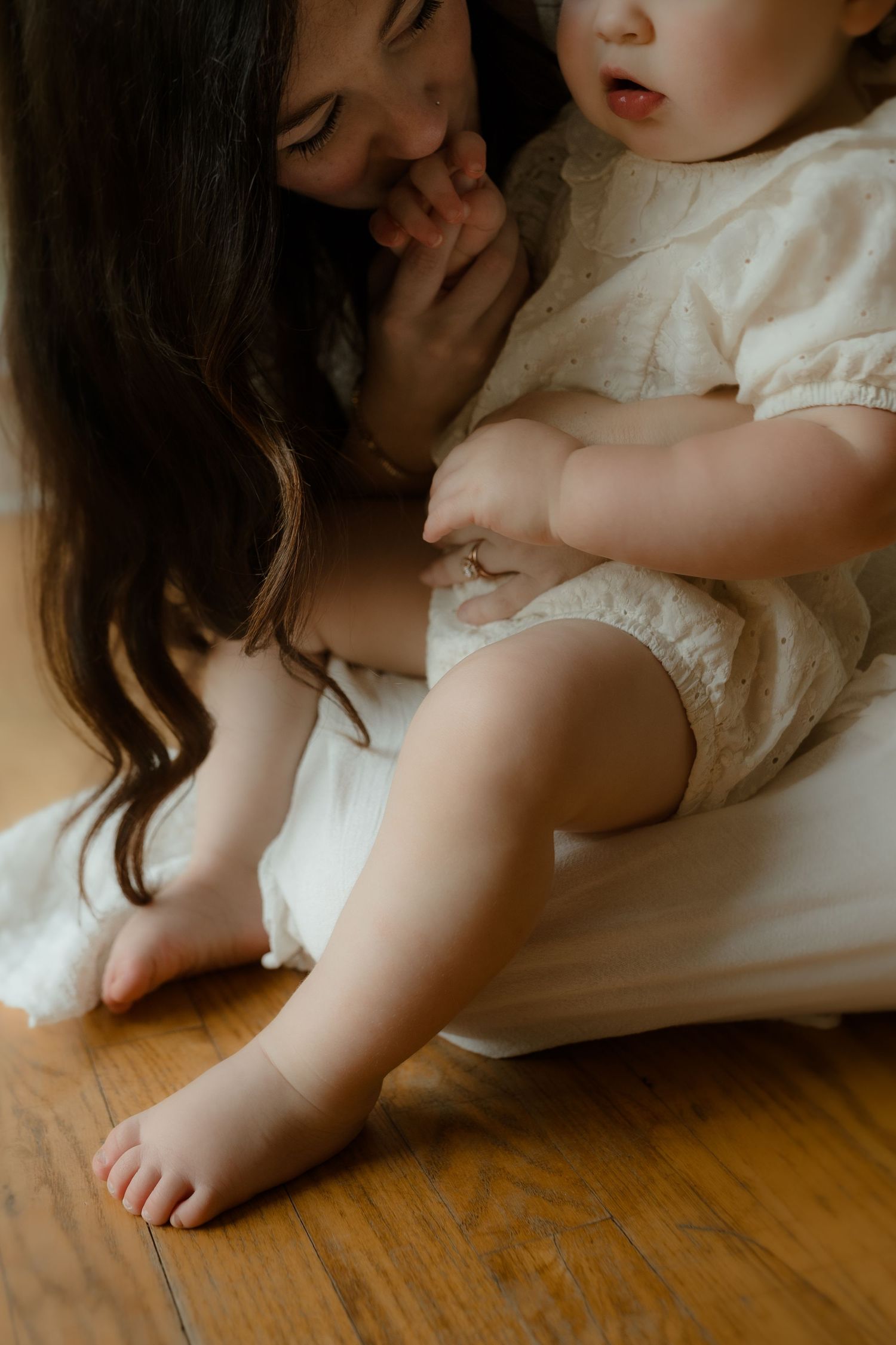 A mother tenderly holds her baby in a cream romper while sitting on a white cushion on a hardwood floor.